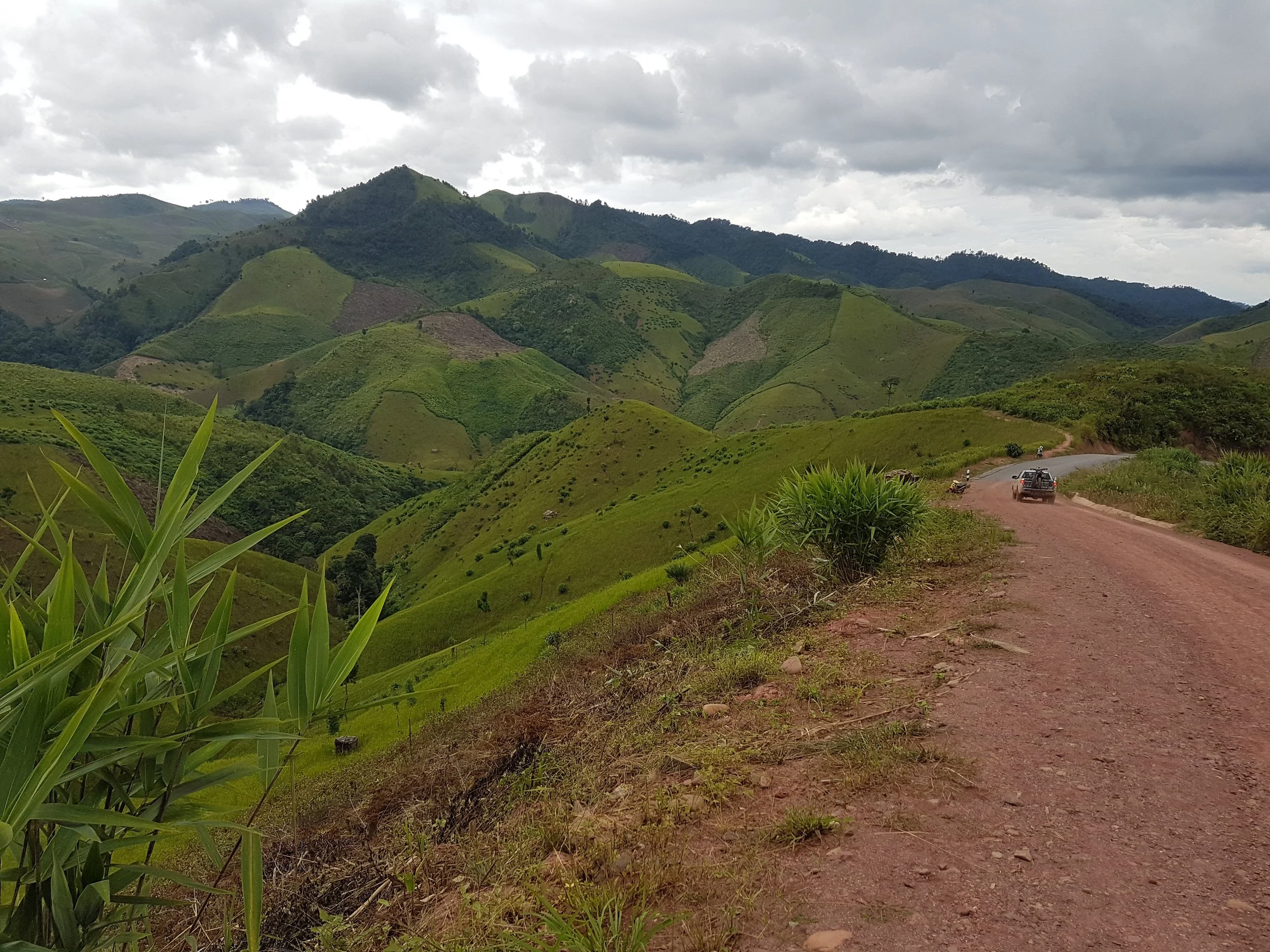 Paysage de montagnes verdoyantes sous un ciel nuageux, avec un chemin de terre où une voiture et une moto roulent.