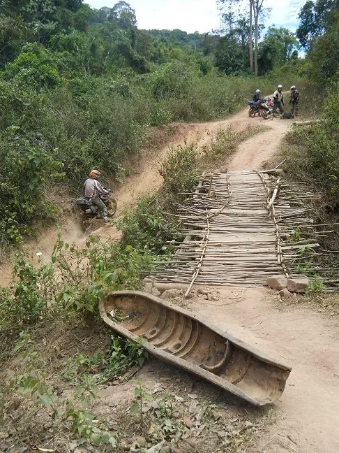 Route en terre avec un pont en bois fait de branches, une barque renversée au premier plan, et des motos avec des personnes portant des casques à l'arrière-plan.