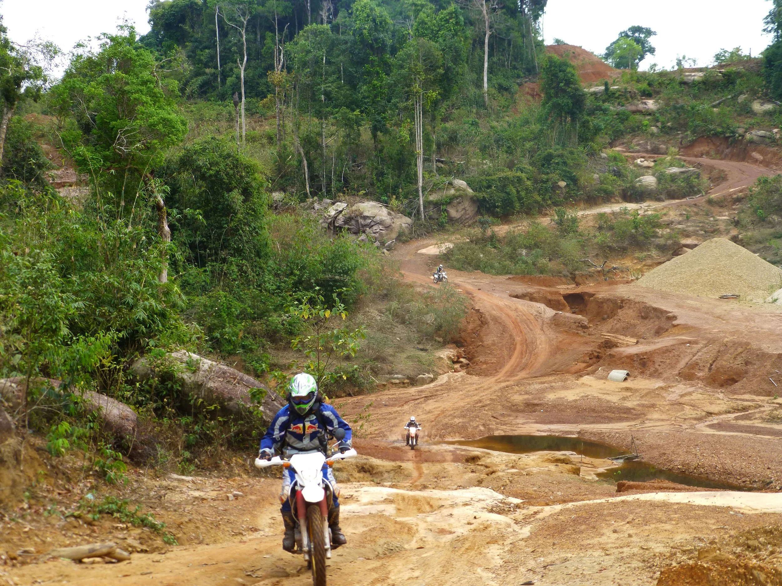 Rallye de motos tout terrain à travers un chemin boueux en zone boisée avec quelques véhicules en arrière-plan. Au Laos.
