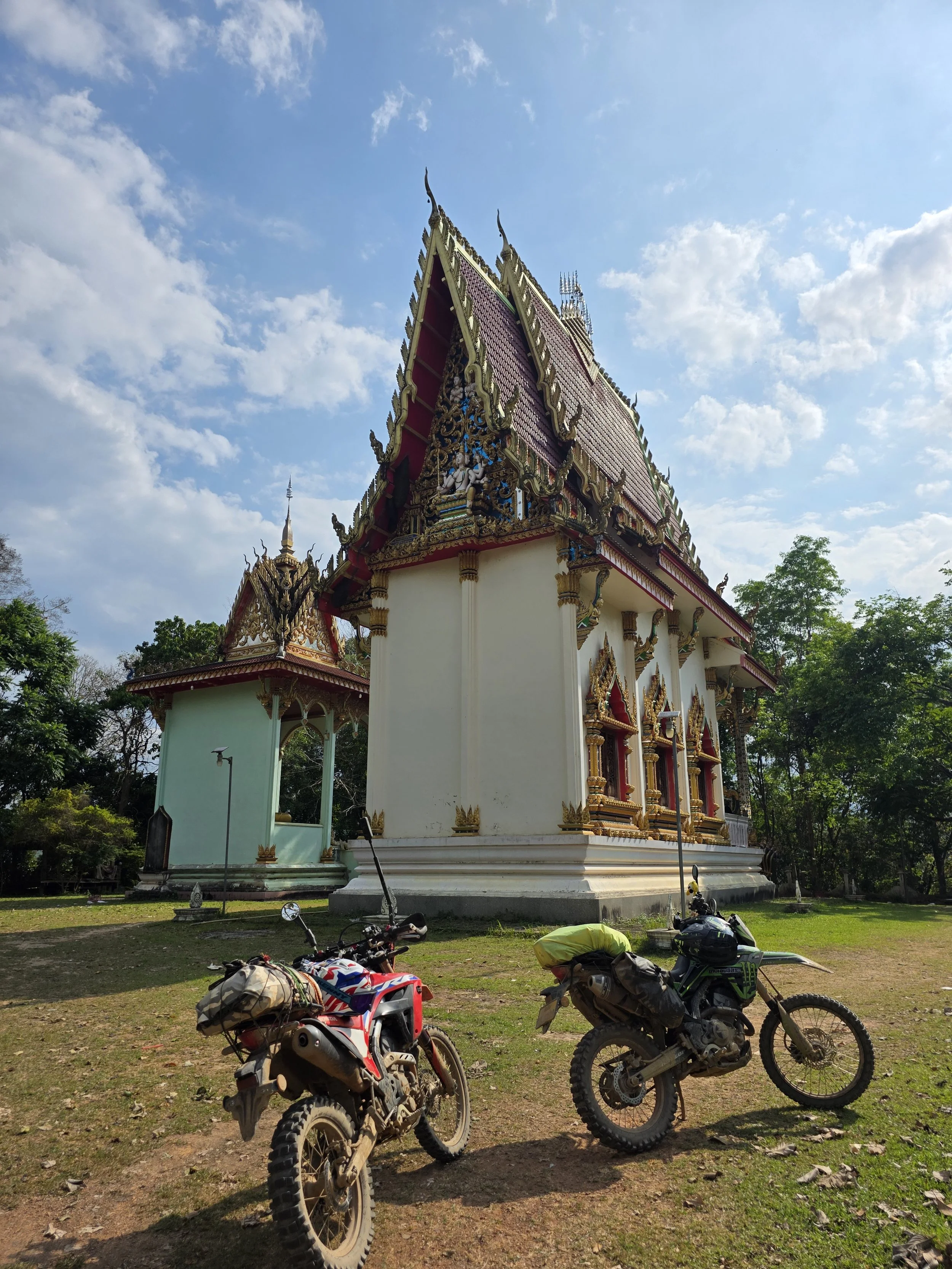 Un temple thaïlandais orné de dorures et de sculptures, entouré d'arbres, avec des motos garées devant.