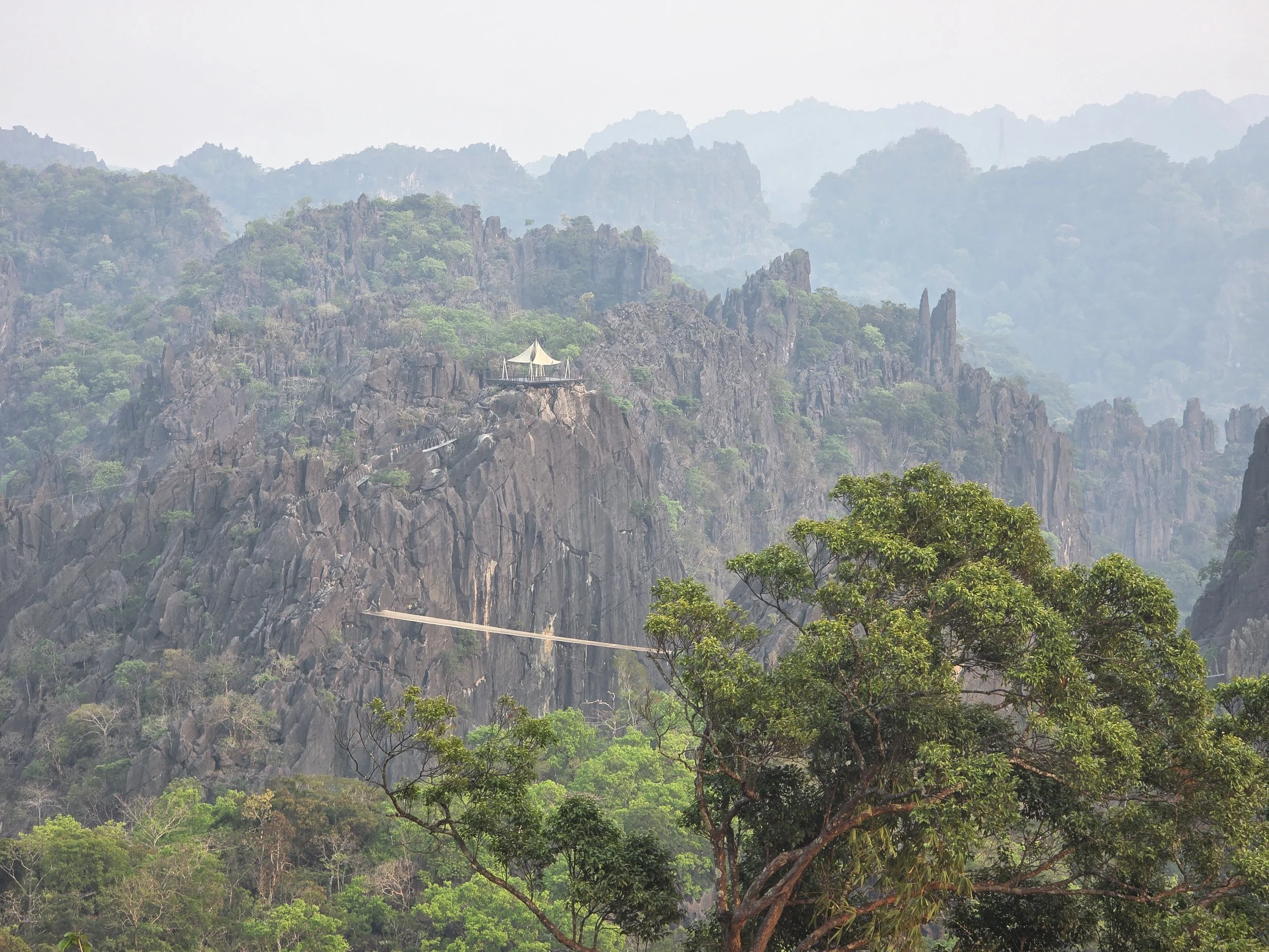 Paysage de montagnes rocheuses avec une végétation dense et un pavillon suspendu avec une tente blanche à proximité, entouré de brouillard ou de brume. Laos