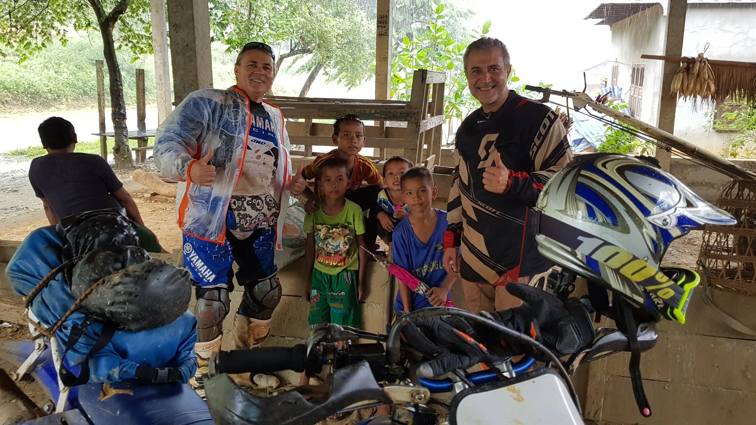 Groupe d'enfants et de deux hommes posant pour une photo dans un environnement rural. Des motos et un casque de moto sont visibles en premier plan.