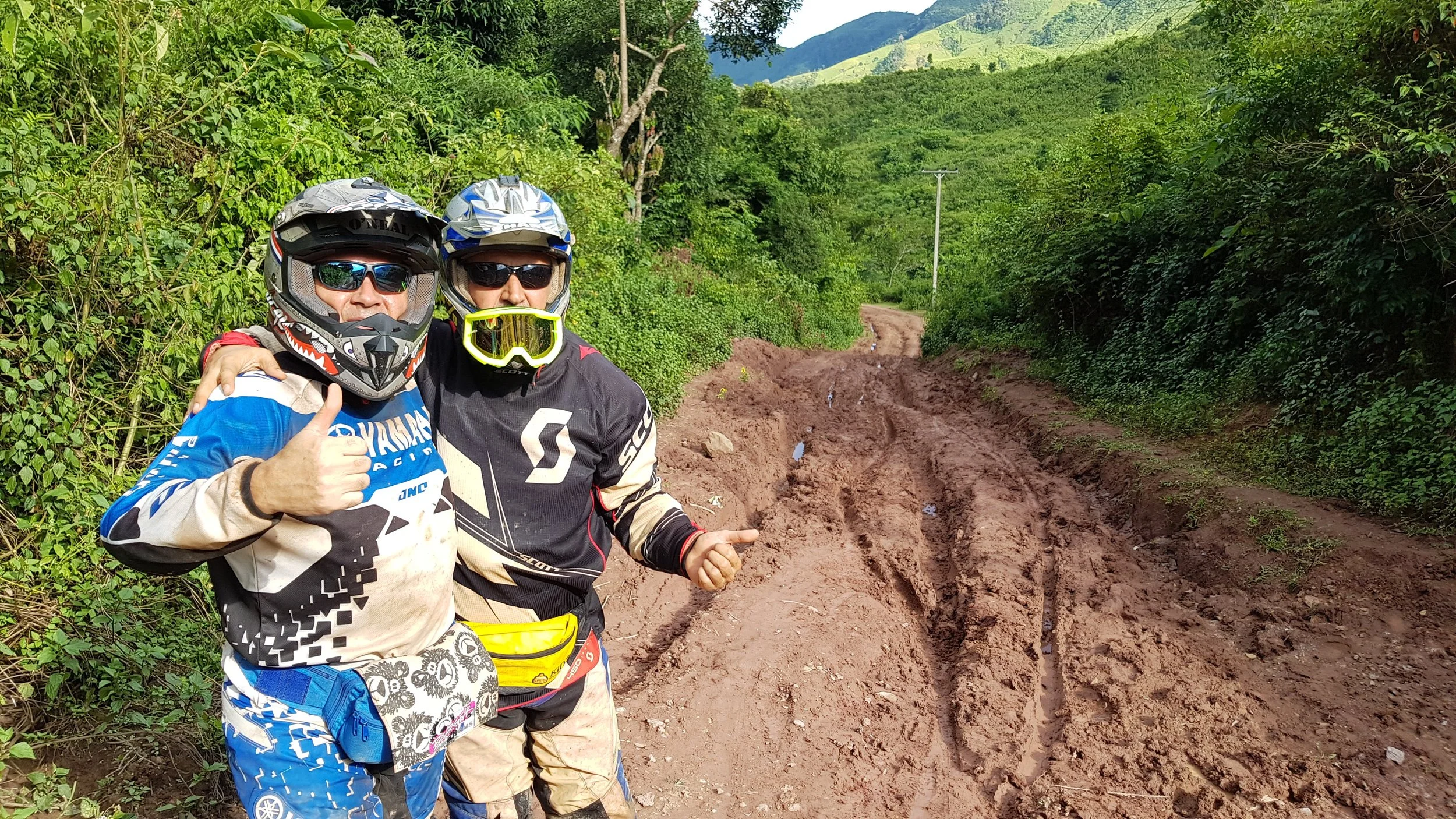 Deux hommes en équipement de motocross posent côte à côte dans un environnement naturel avec de la verdure et des montagnes en arrière-plan. Ils portent des casques, des lunettes de protection et des vêtements de motocross, l'un donnant un coup de po