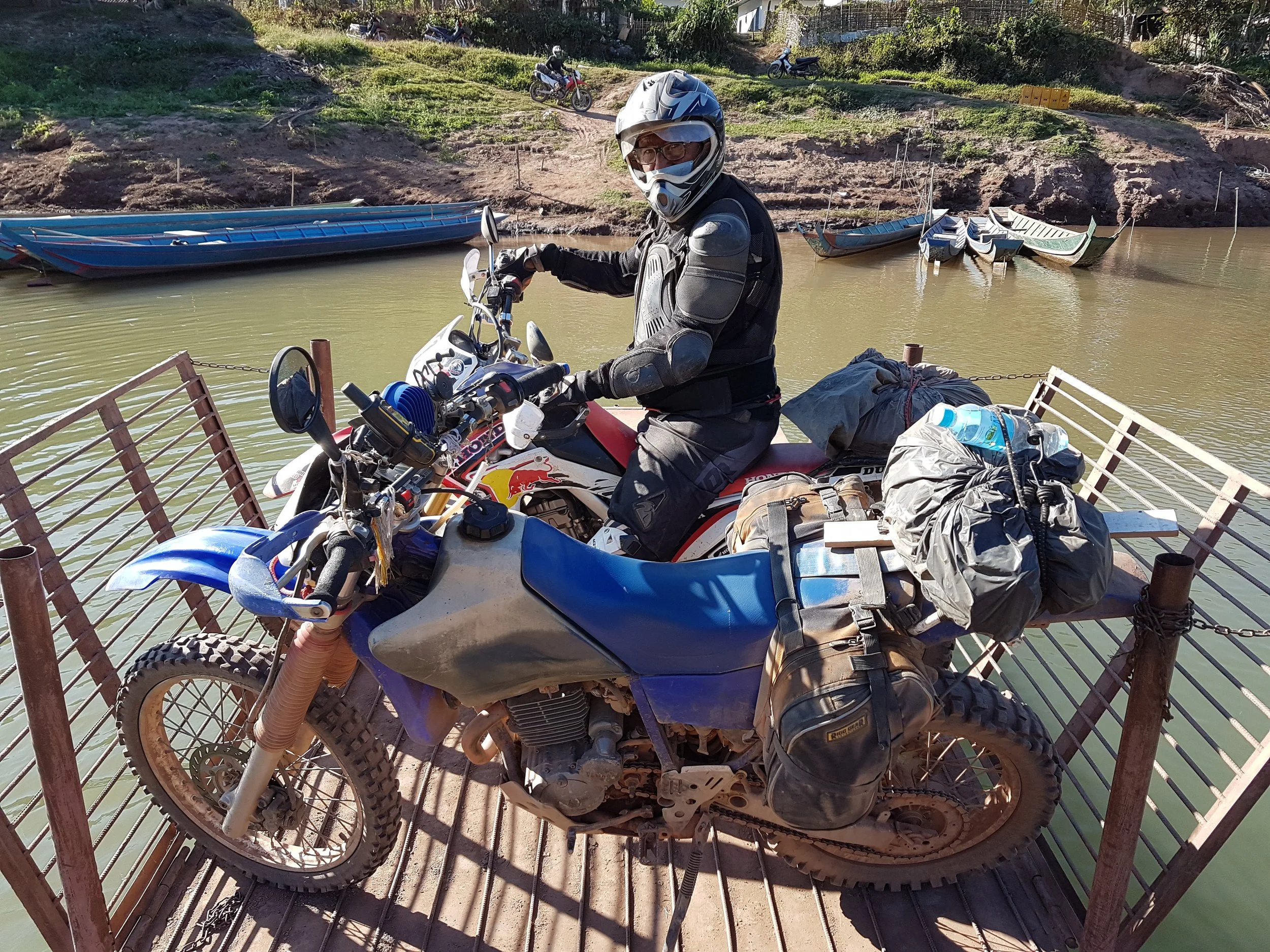 Un homme portant un casque et des équipements de moto tout-terrain, assis sur une moto dans une barge en bois, avec des bateaux en bois à l'arrière, le long d'une rivière ou d'un lac, entouré d'une nature verdoyante.
