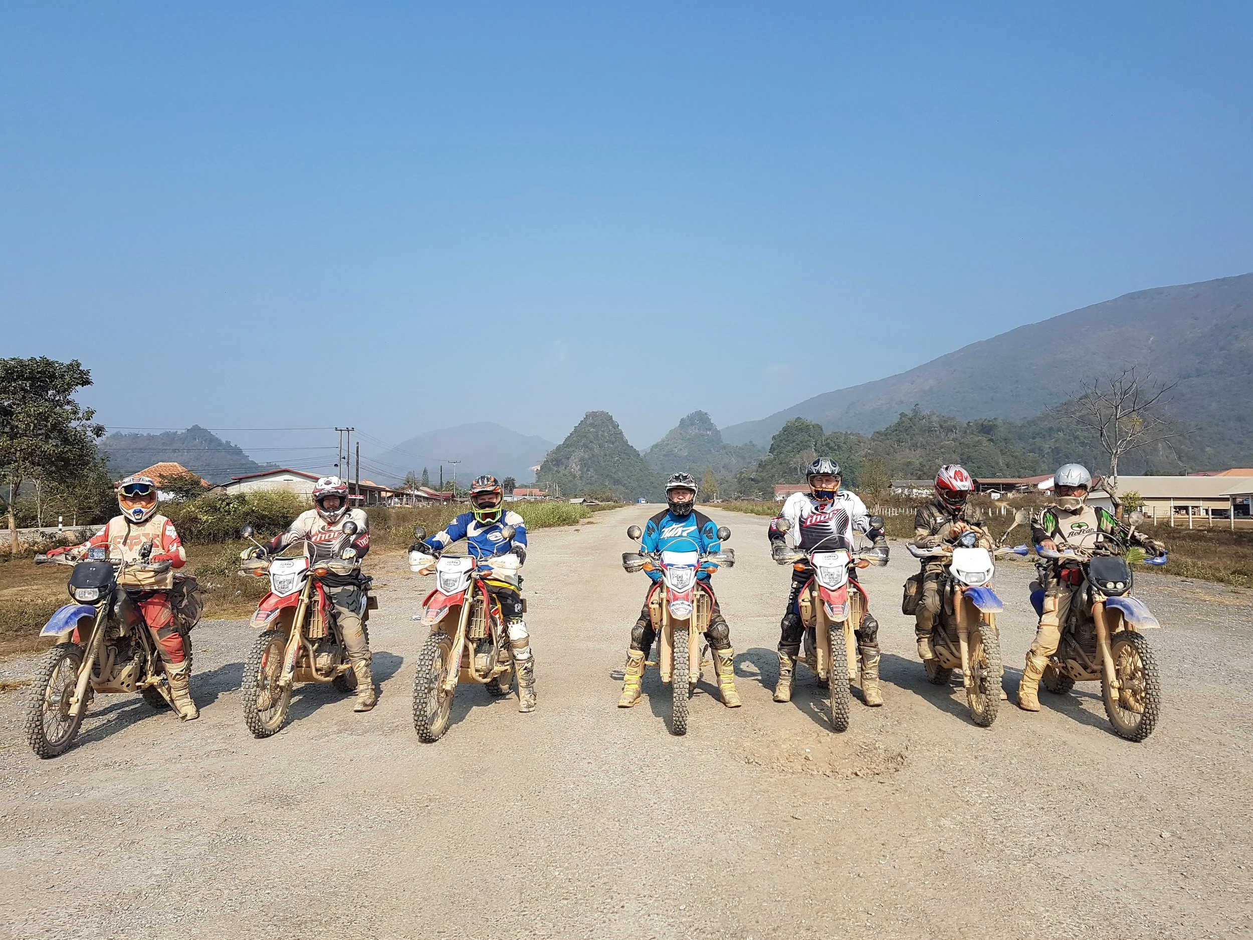 Groupe de huit motards en équipement de protection, alignés en ligne, sur des motos tout-terrain, dans une zone champêtre avec des montagnes en arrière-plan, sous un ciel clair.