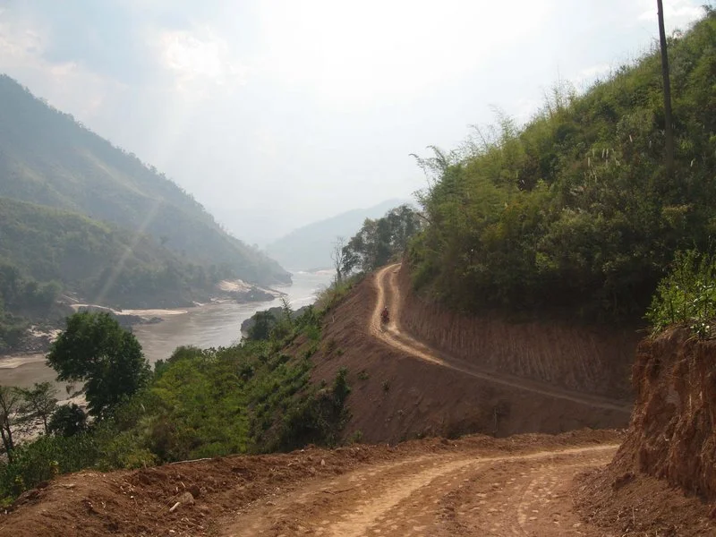 Une route de terre sinueuse longe une rivière dans une vallée entourée de montagnes verdoyantes, par une journée ensoleillée avec quelques nuages.