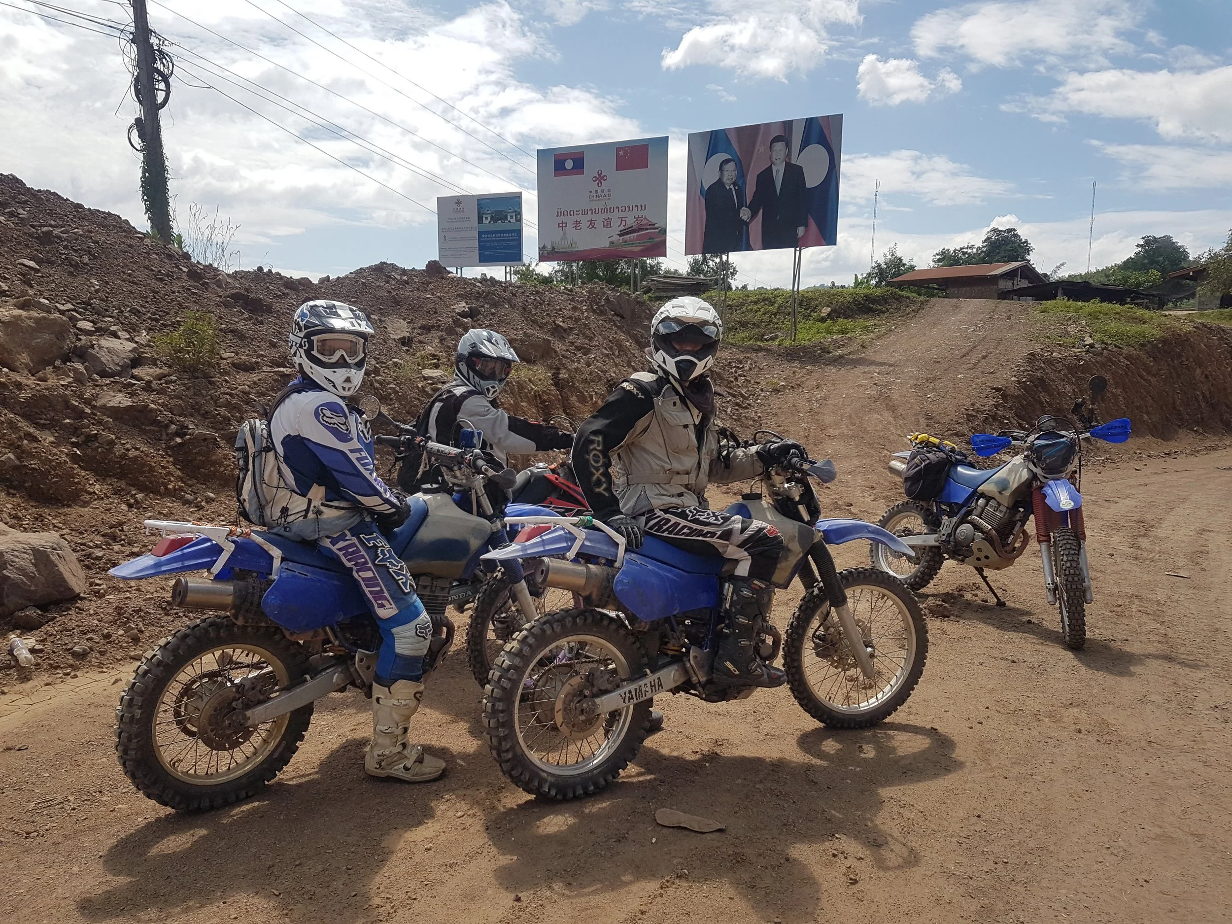 Groupe de motards avec des casques et des vêtements de protection, posant pour une photo sur une route en terre, avec des panneaux publicitaires en arrière-plan, dont un montrant une réunion entre deux hommes en costume. Le ciel est partiellement nua
