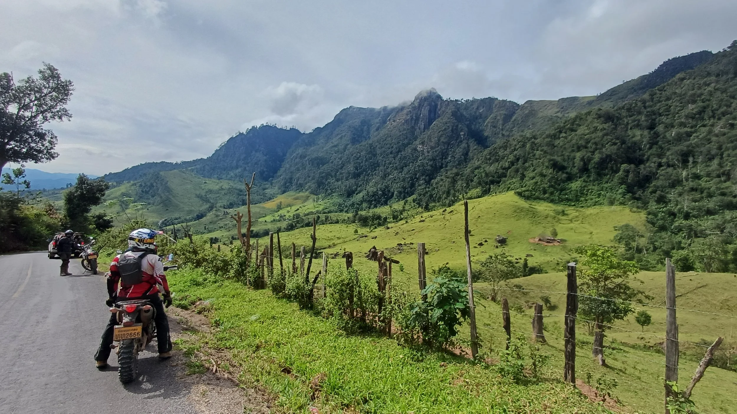 Des motards arrêtés sur une route de campagne entourée de collines verdoyantes et de montagnes en arrière-plan, sous un ciel nuageux.