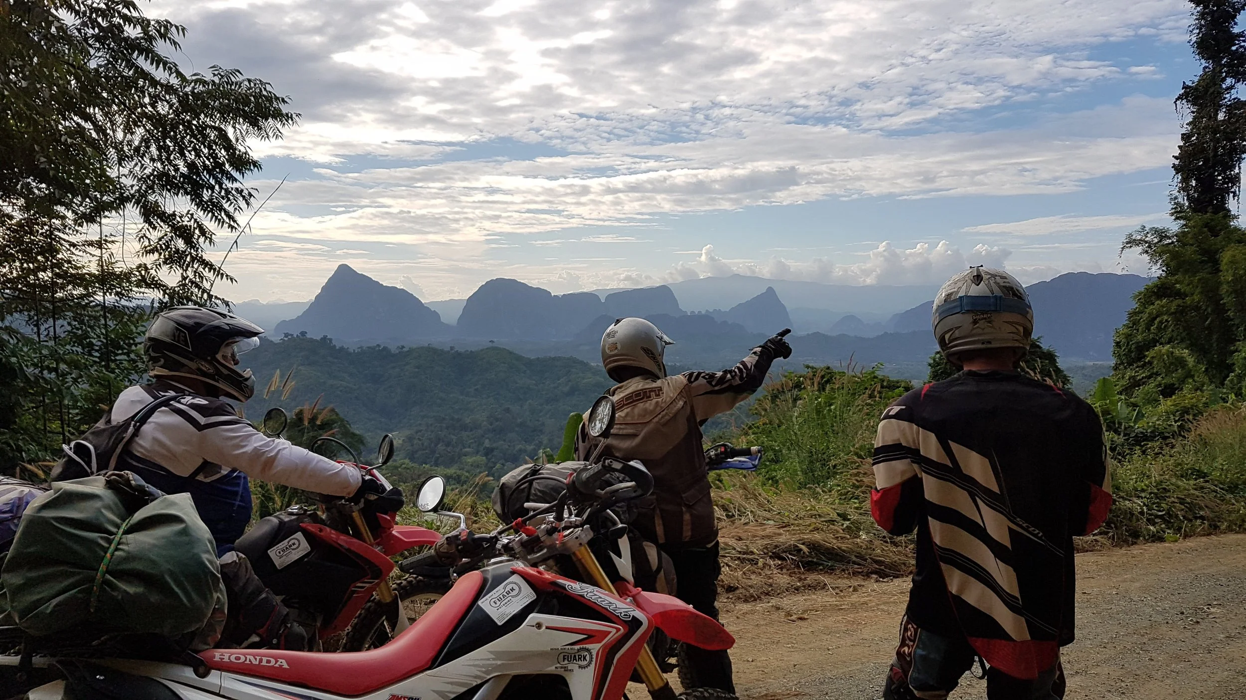 Trois motards regardant un paysage montagneux avec des montagnes et un ciel nuageux, dans un environnement forestier ou rural.