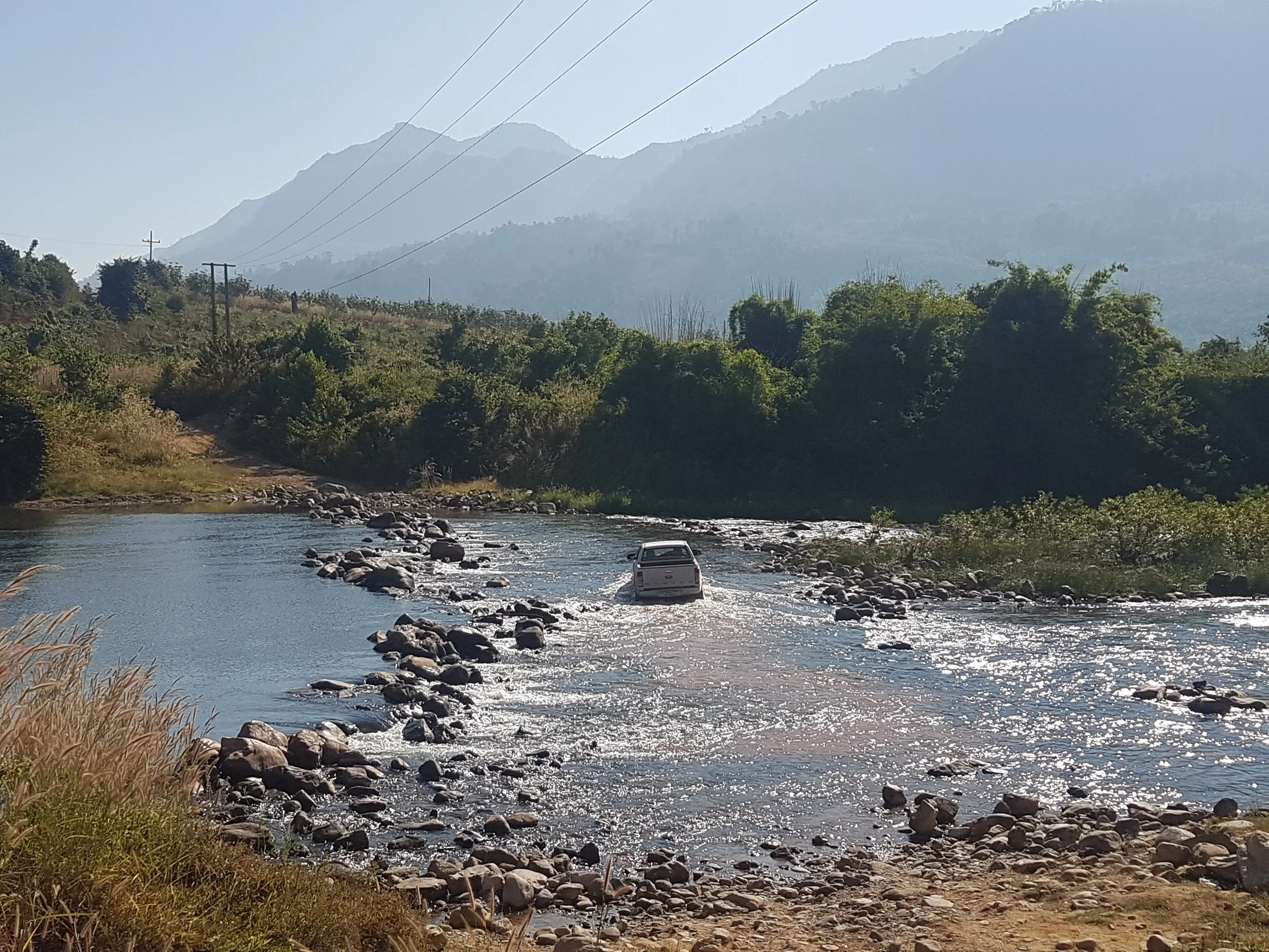 Une rivière coule à travers un paysage de montagnes verdoyantes avec des arbres et des buissons, un véhicule blanc est visible dans l'eau, des lignes électriques parcourent le ciel clair.
