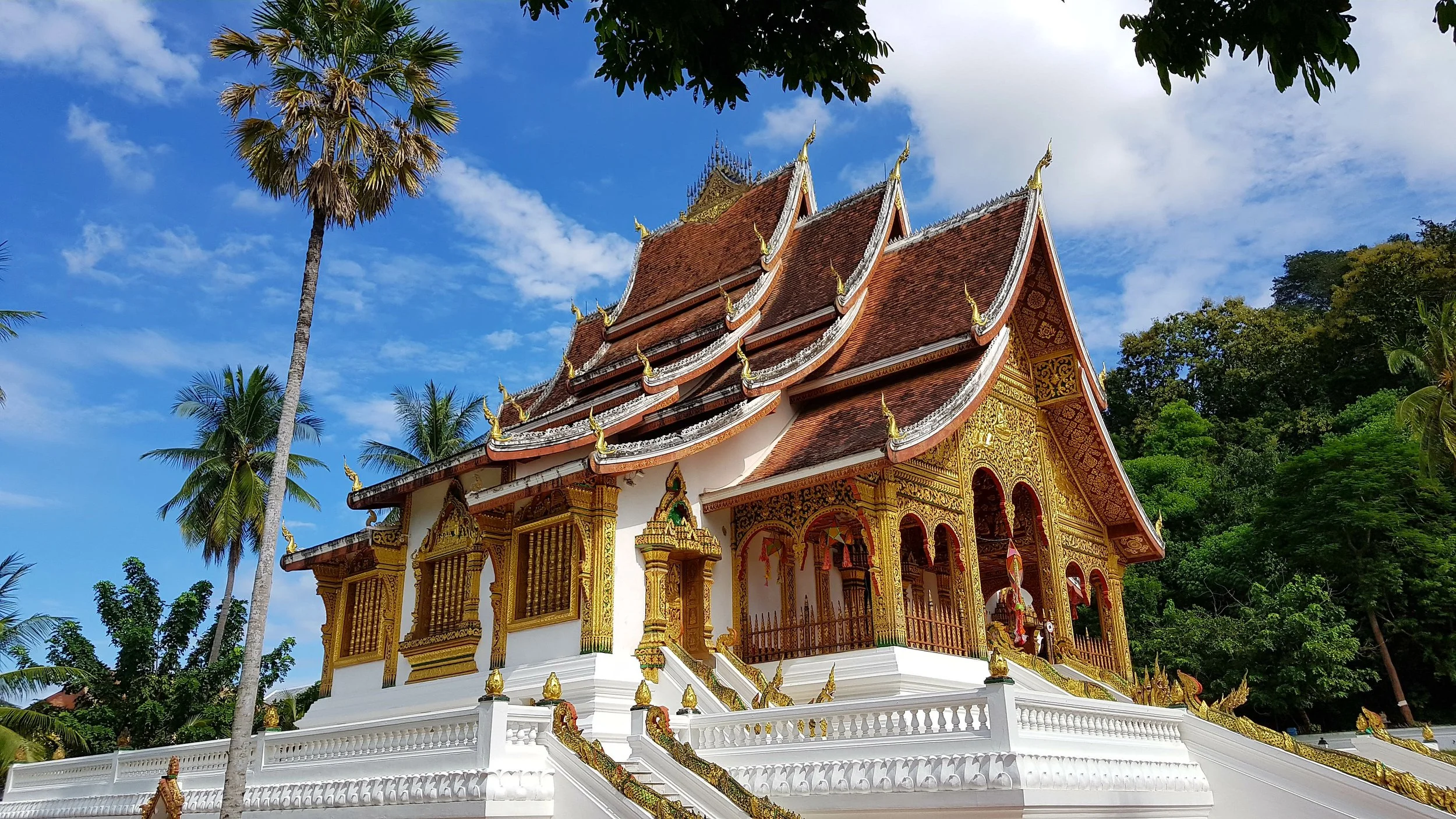 Un temple thaïlandais traditionnel avec un toit en pente orné de détails dorés, entouré de végétation tropicale et de palmiers, sous un ciel bleu avec quelques nuages.