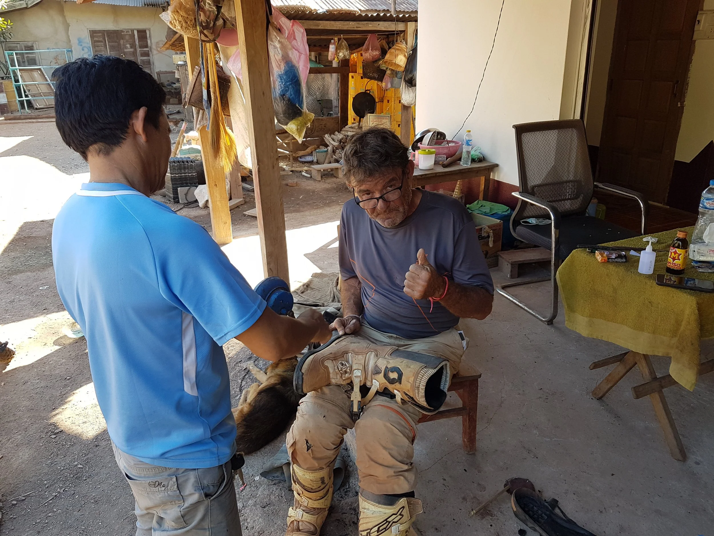 Deux hommes assistent un chien portant des bottes de travail, à l'extérieur dans une zone avec des meubles et des outils. Un homme à gauche porte une chemise bleue, l'autre à droite porte des lunettes et un t-shirt gris.