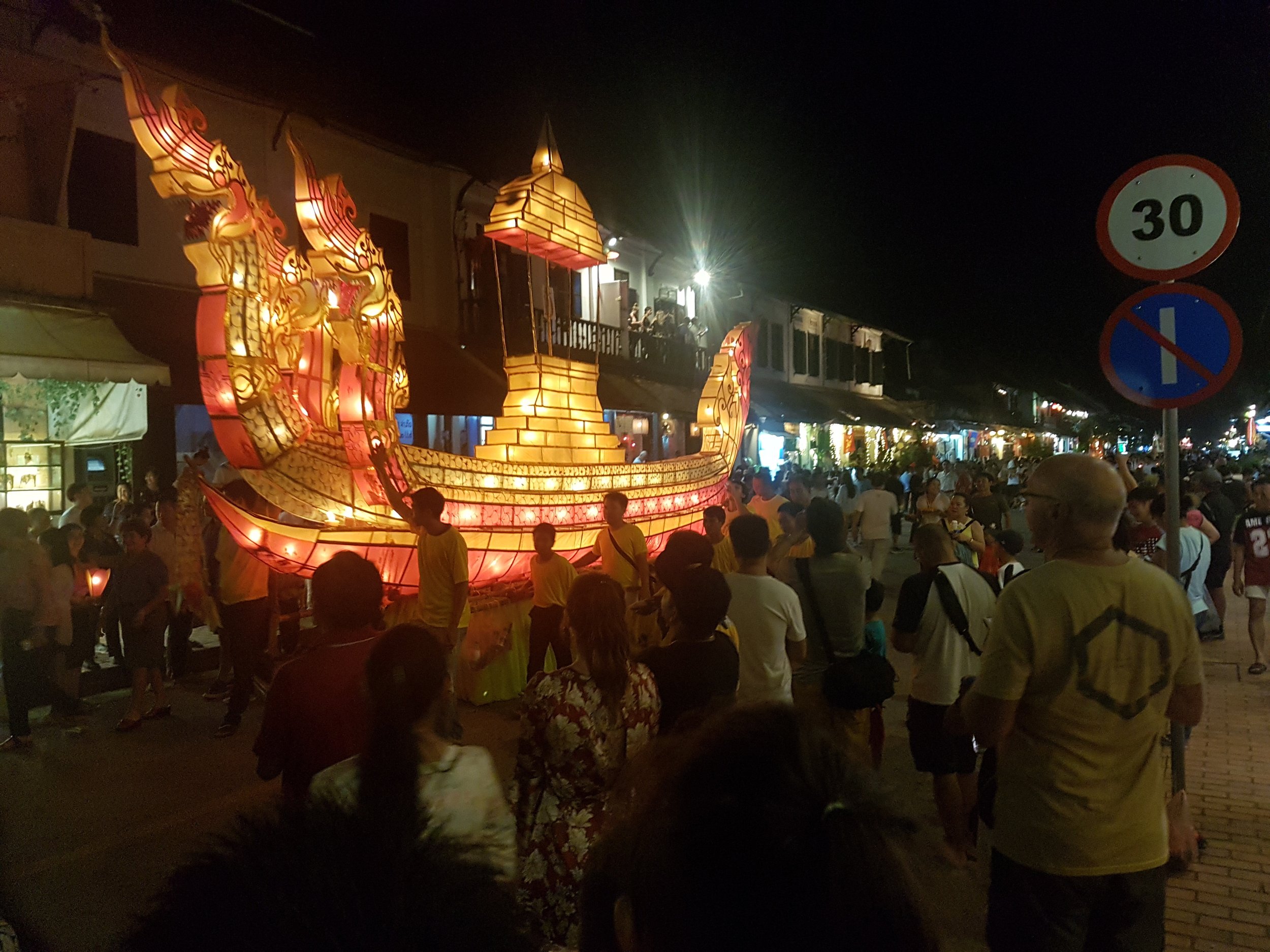 Une parade nocturne avec une grande structure illuminée en forme de bateau avec des dragons, entourée d'une foule de spectateurs. Des décorations lumineuses et des enseignes de circulation sont visibles.