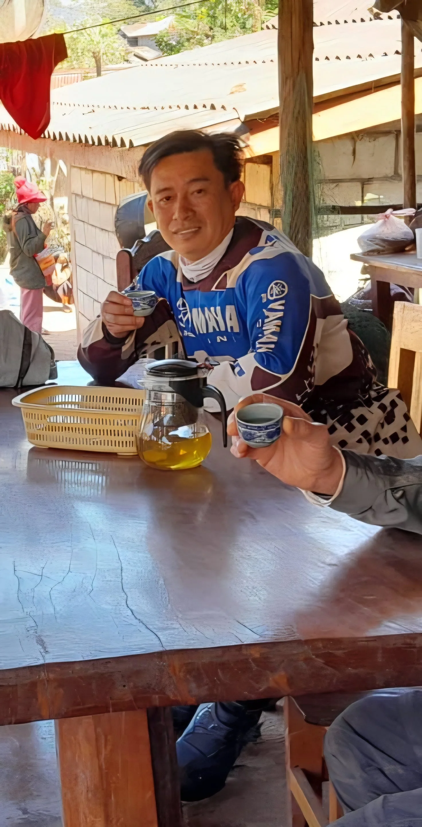 Deux personnes trinquent avec des tasses à thé ou à café en bois, assises à une table en bois dans un environnement rustique, avec un homme en veste de MotoGP et une femme en arrière-plan portant un chapeau rouge. Au Laos.