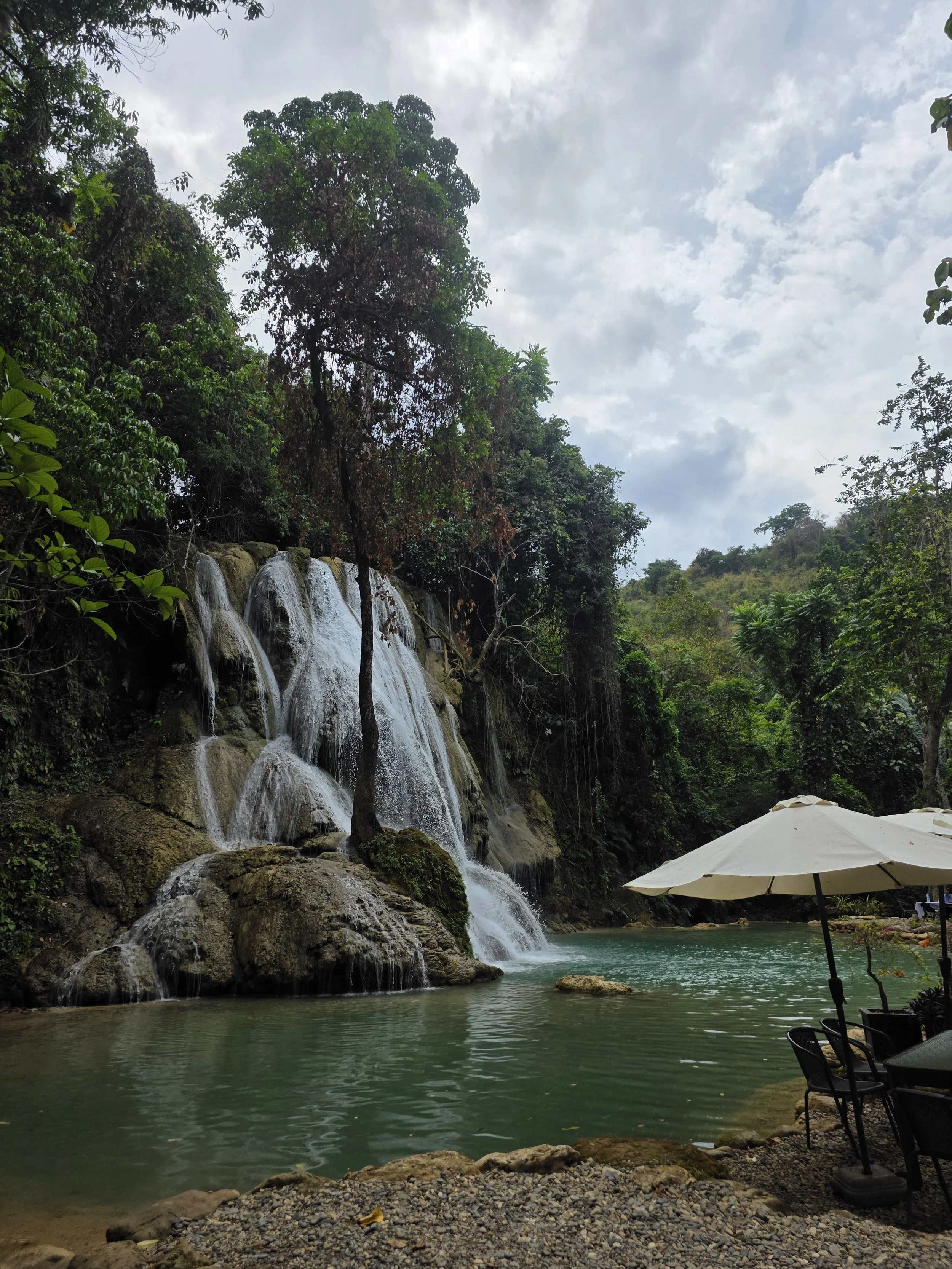 Une chute d'eau dans une forêt tropicale, avec une piscine naturelle en bas, une parasol blanc et des chaises, ambiance paisible et naturelle.