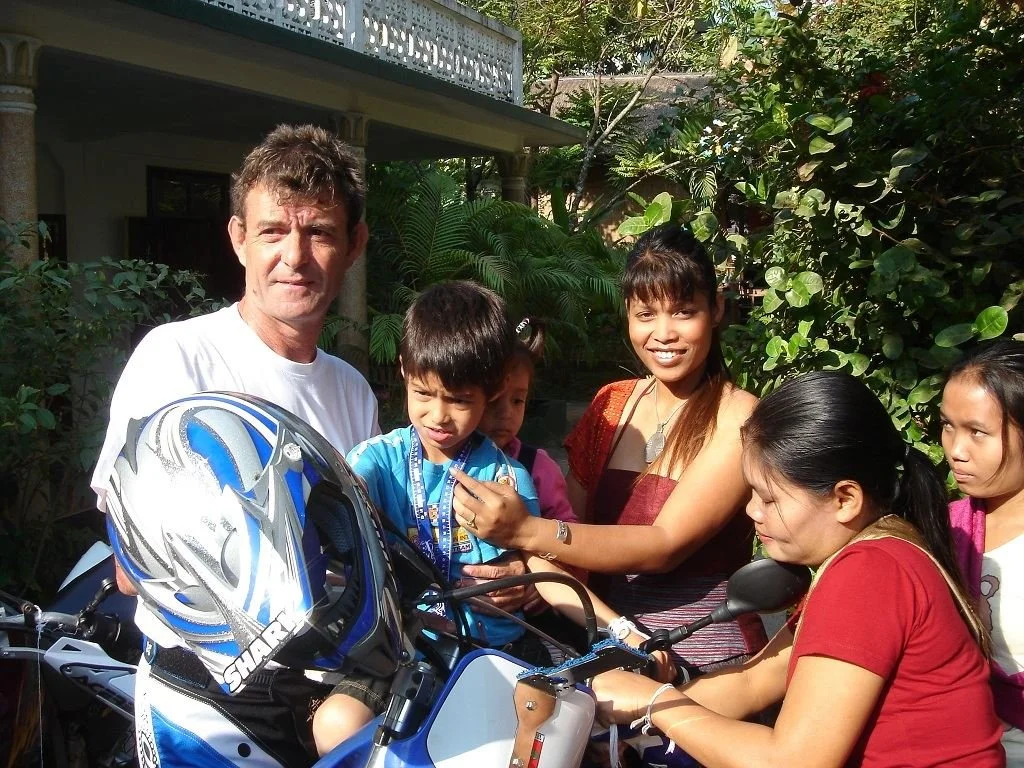 Un groupe de personnes, dont un homme, une femme et plusieurs enfants, posant près d'une moto, en extérieur avec des plantes et une maison en arrière-plan.
