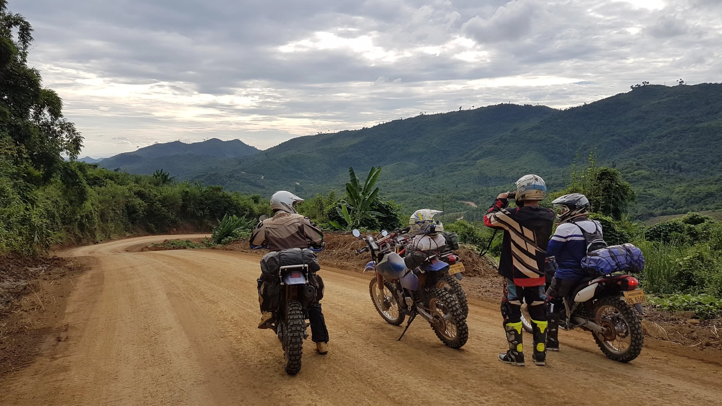 Groupe de motards avec casques et sacoches sur une route de campagne en lacets, entourée de végétation et de montagnes sous un ciel nuageux.