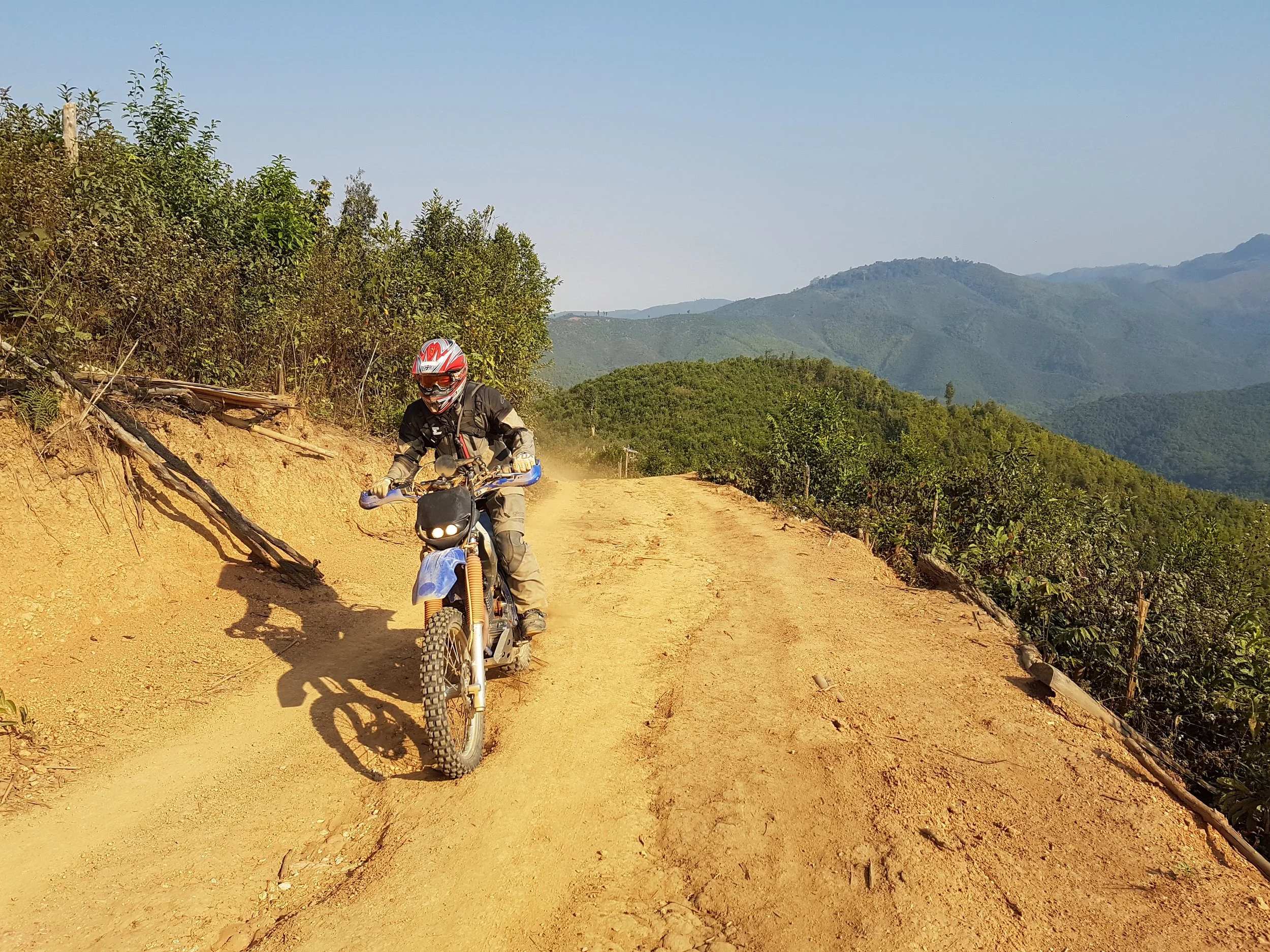 Une personne portant un casque de moto et des vêtements de protection, roulant sur un chemin de terre dans un paysage montagneux avec des arbres et un ciel dégagé.
