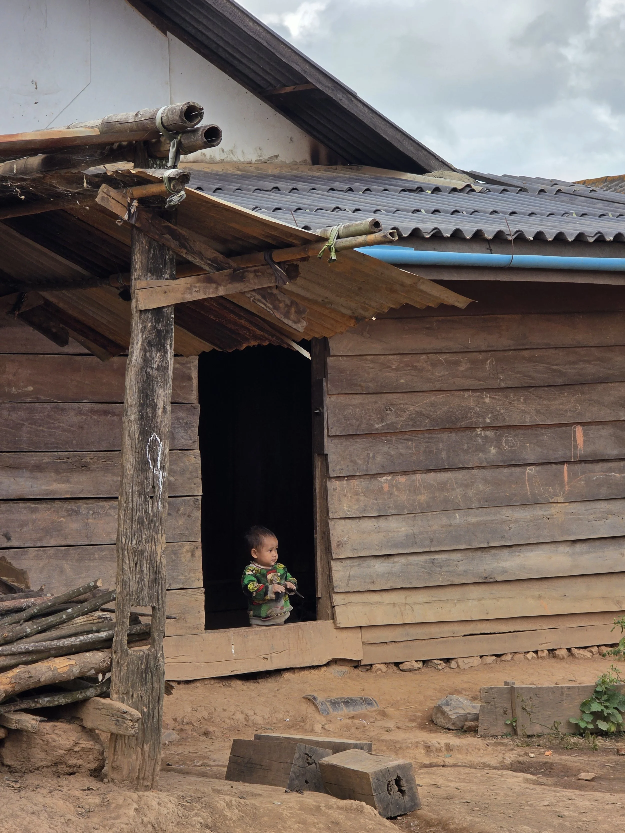 Un enfant debout dans l'entrée d'une maison en bois, tenant un objet dans ses mains, sous un ciel nuageux.