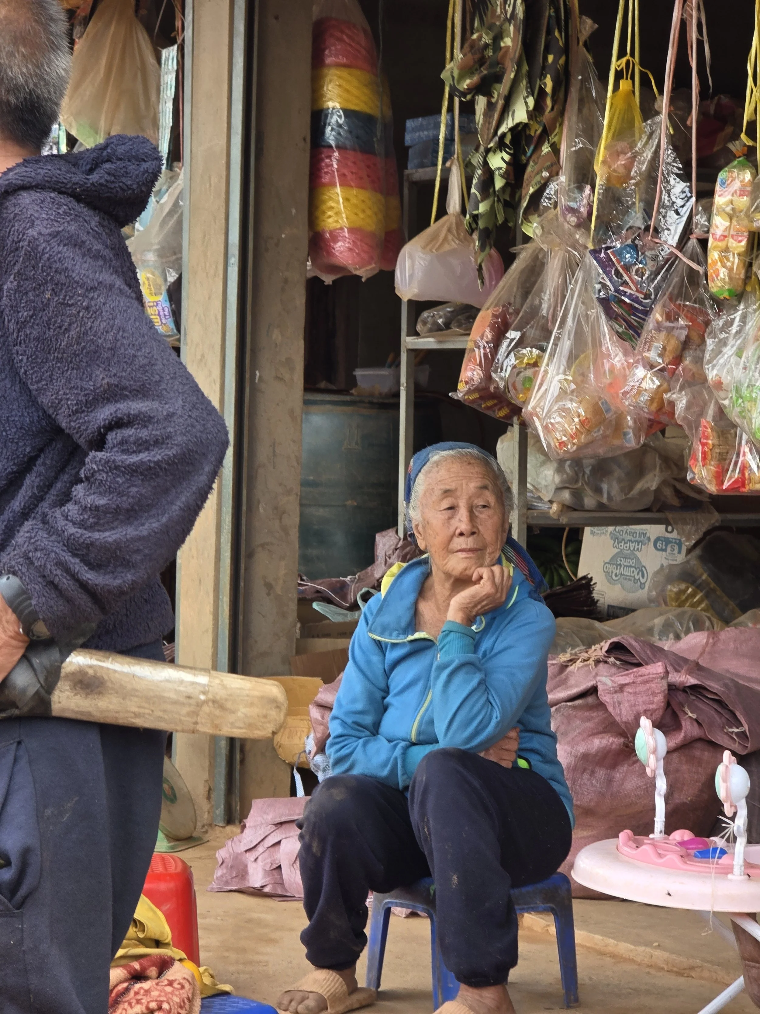 Une femme âgée assise sur une petite chaise bleue dans un magasin ou marché, entourée d'articles divers comme des sacs en plastique avec des produits, avec des personnes autour.