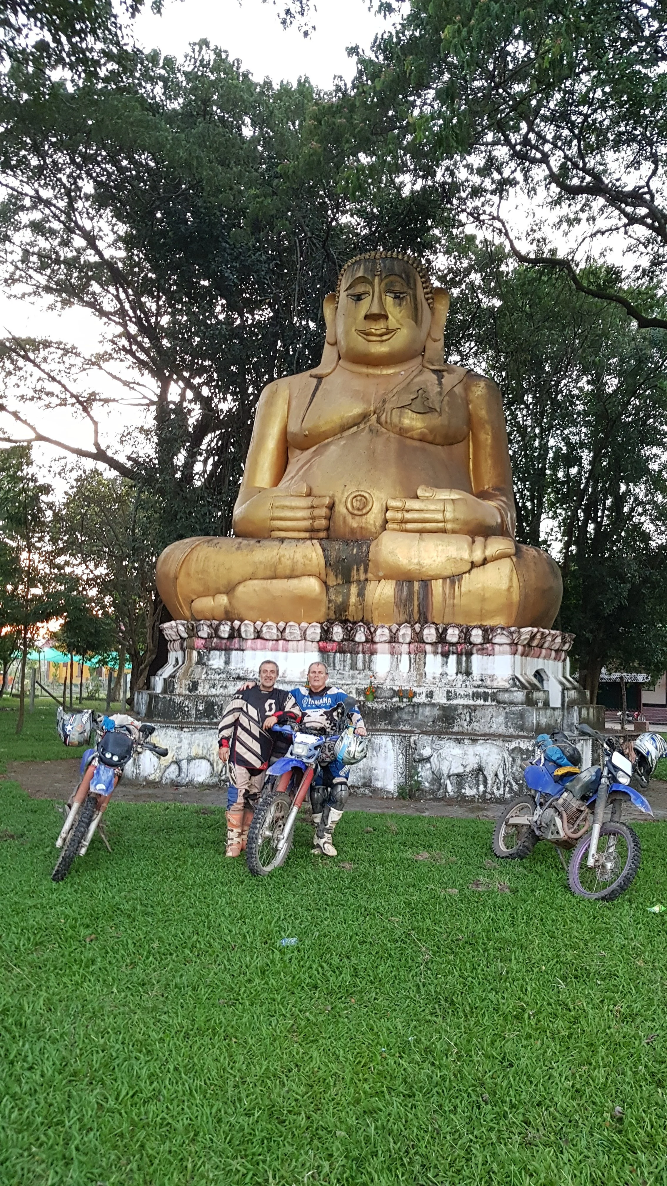Gros symbole religieux doré représentant une figure assise méditative, entouré d'arbres verts. Il y a deux hommes en moto devant la statue, près de plusieurs autres motos.