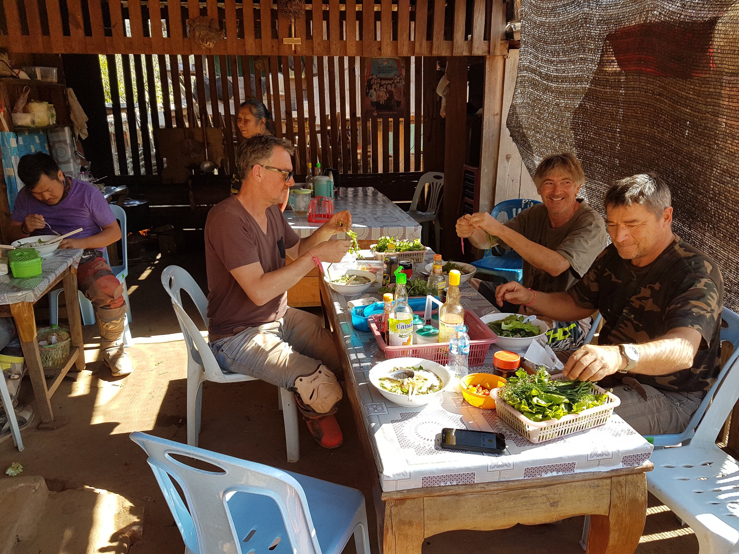 Un groupe de cinq personnes mangeant à une table en plein air, avec des plats asiatiques et des boissons. L'ambiance semble conviviale et détendue, dans un environnement rustique en bois.