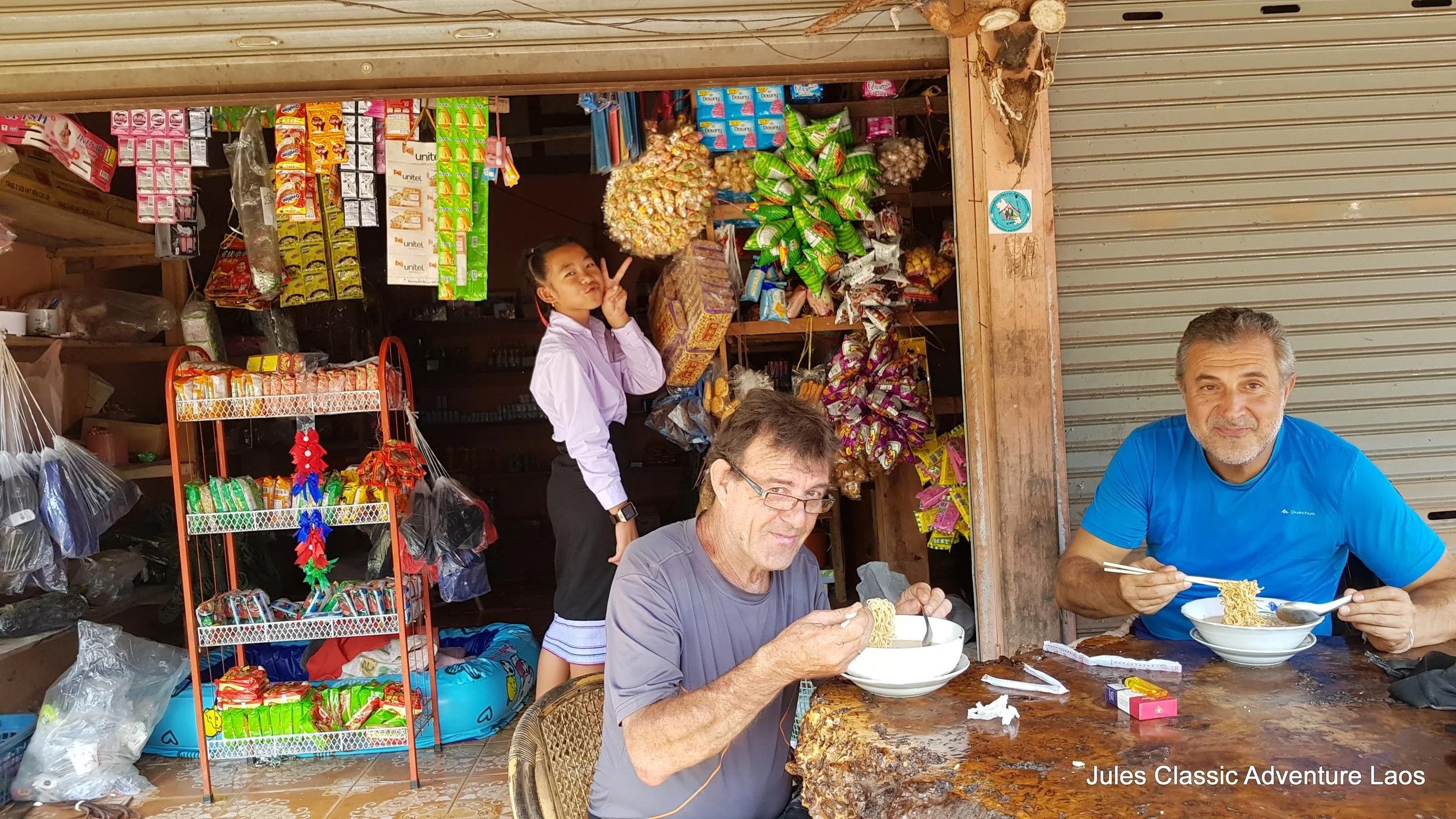 Trois personnes mangent des nouilles dans une cuisine de rue, avec une boutique de snacks colorés en arrière-plan. Lorsqu'une jeune femme fait un signe de paix à l'arrière-plan. La scène se déroule probablement à Laos.