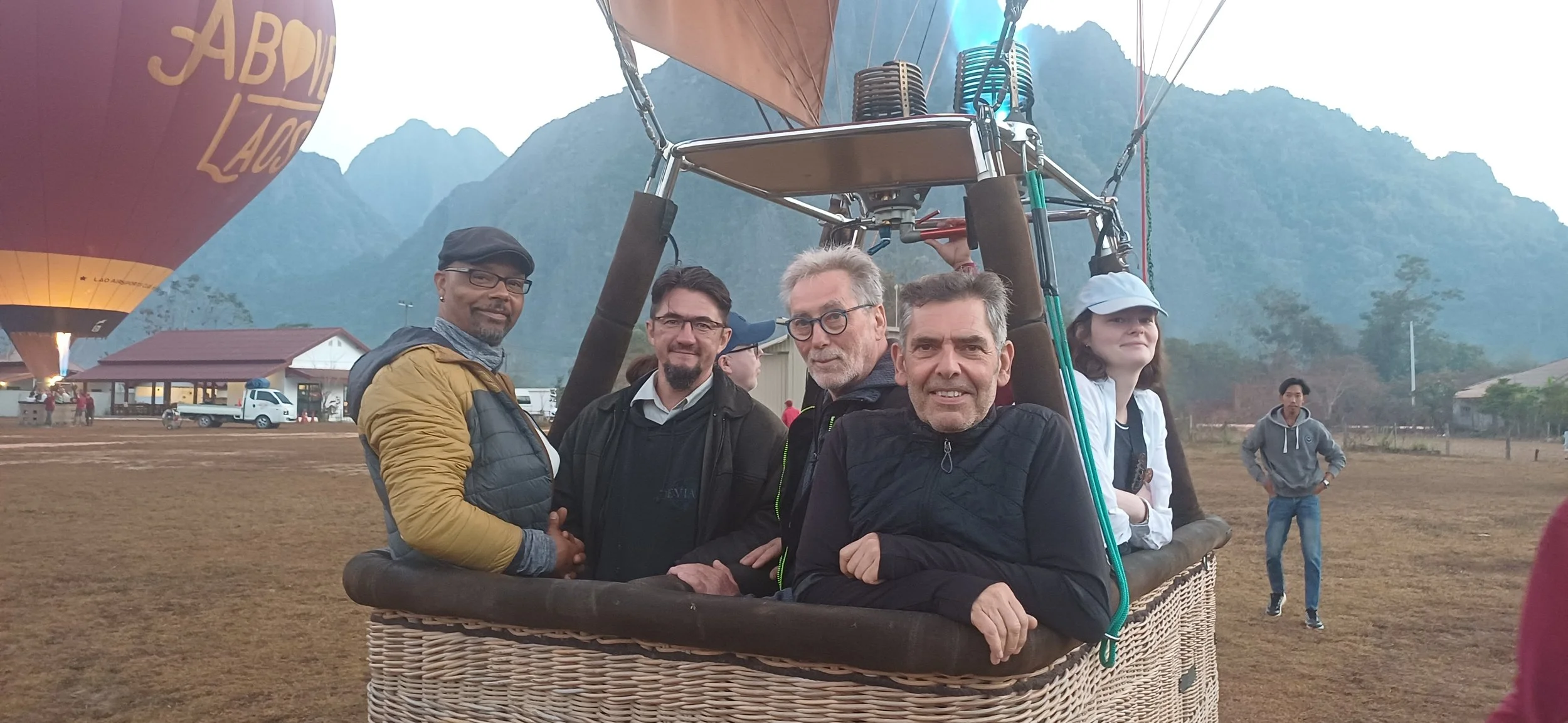 Groupe de personnes dans une nacelle de montgolfière, avec un paysage montagneux en arrière-plan. Un ballon de montgolfière est visible à gauche.