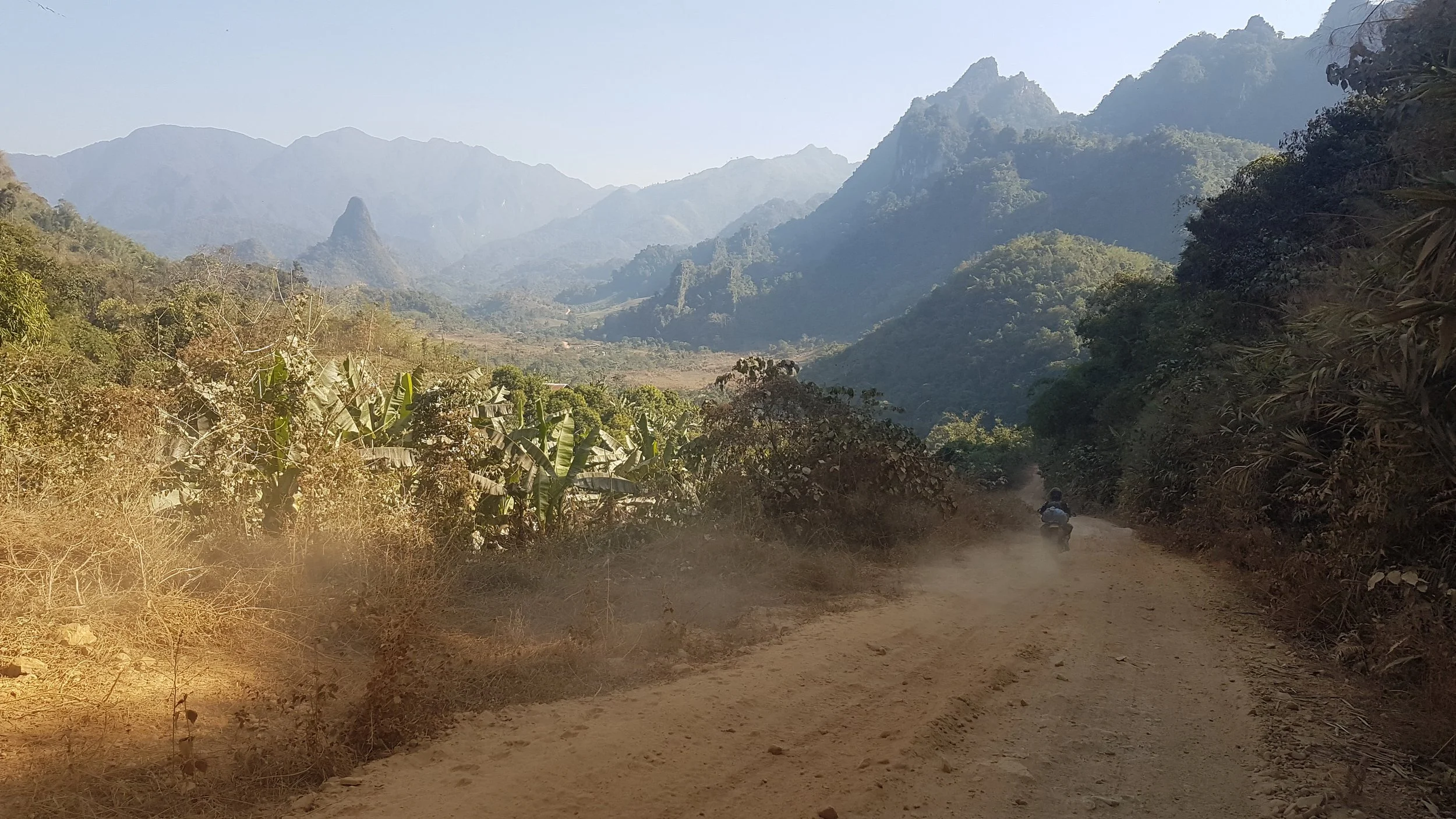 Vue d'une route en terre dans une vallée entourée de montagnes verdoyantes, avec un véhicule motorisé roulant sur le chemin et une végétation dense partout.