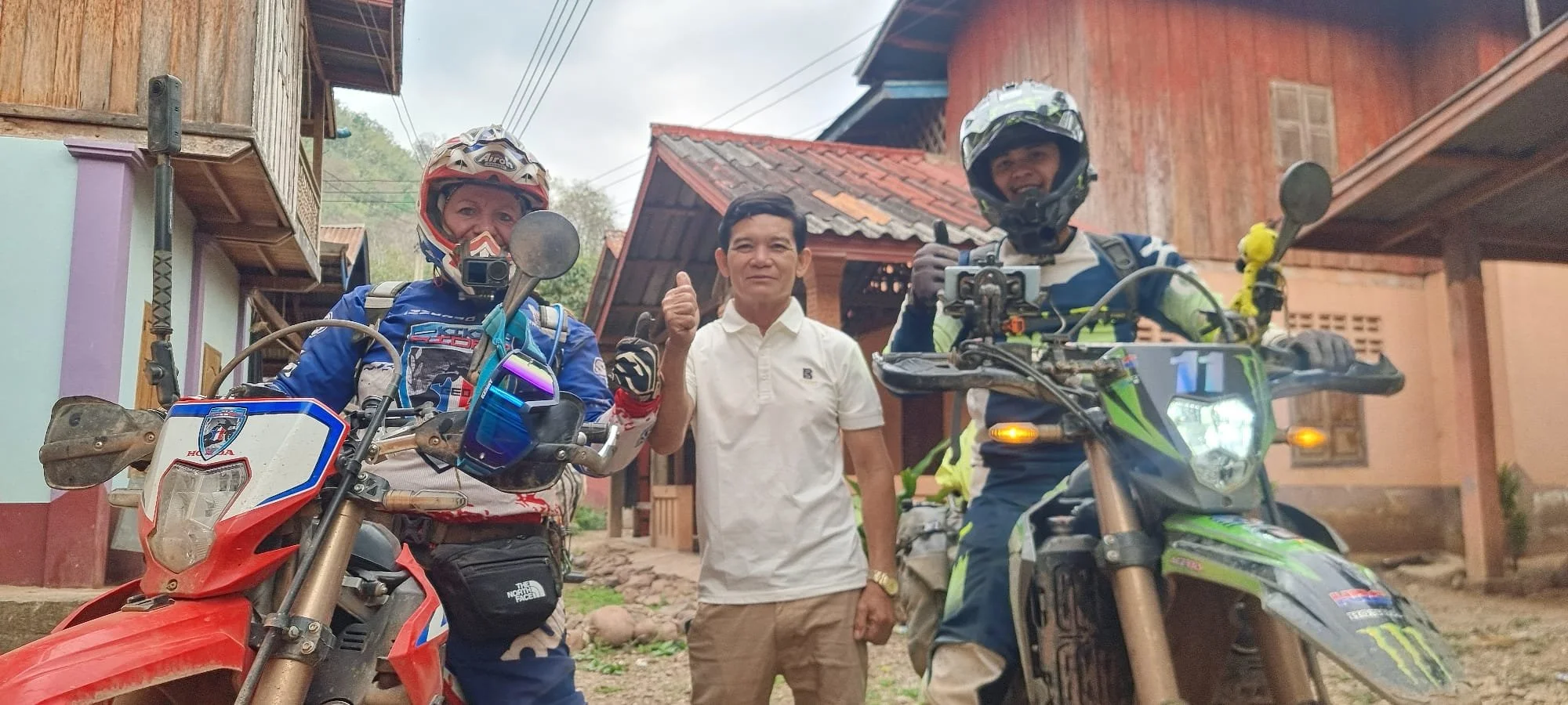 Trois personnes, deux motards en équipement et un homme en chemise, posent ensemble devant des maisons en bois, avec des motos tout-terrain.