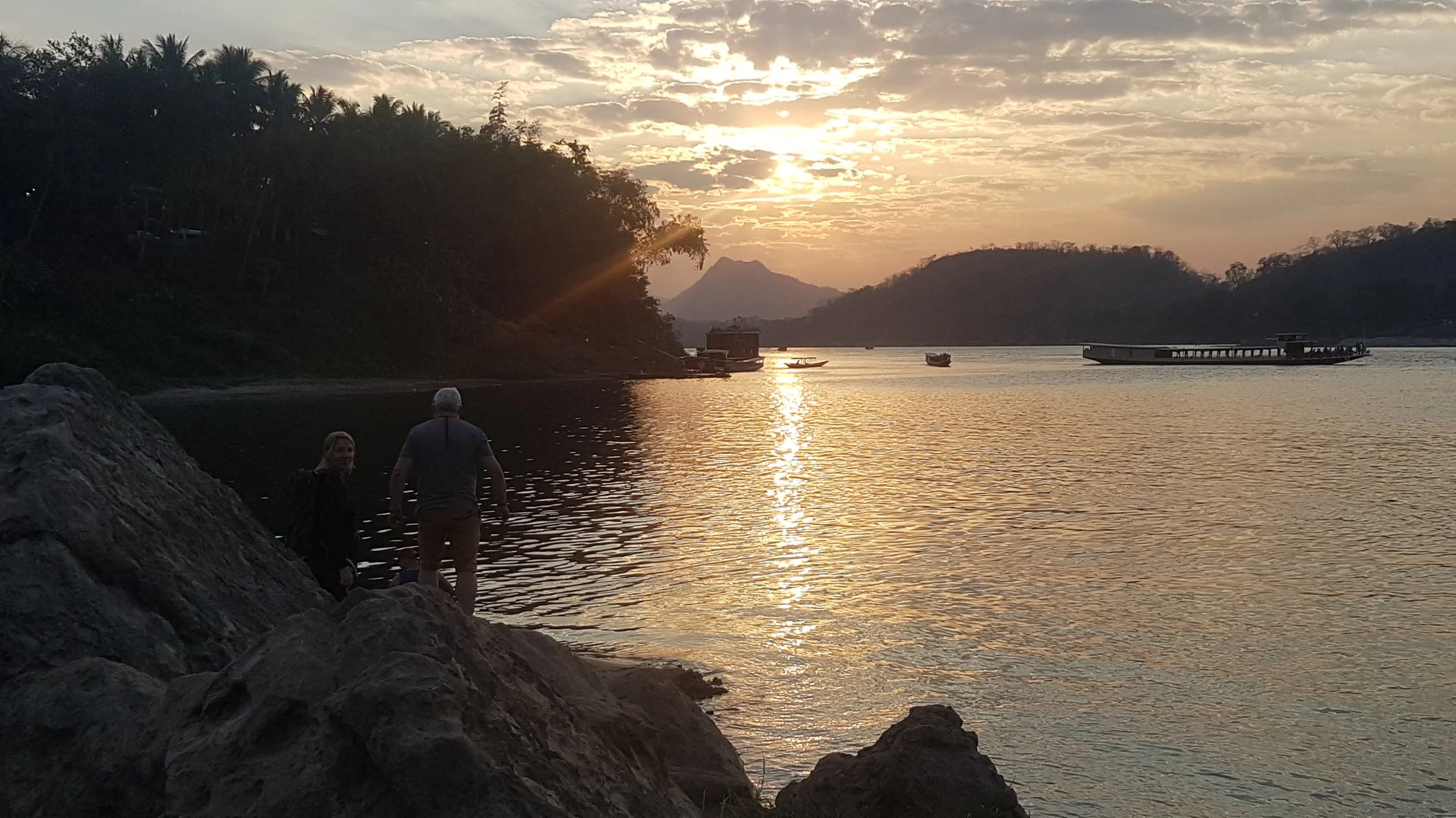 Coucher de soleil sur une rivière avec des montagnes en arrière-plan, des bateaux à quai et deux personnes à proximité des rochers au bord de l'eau.