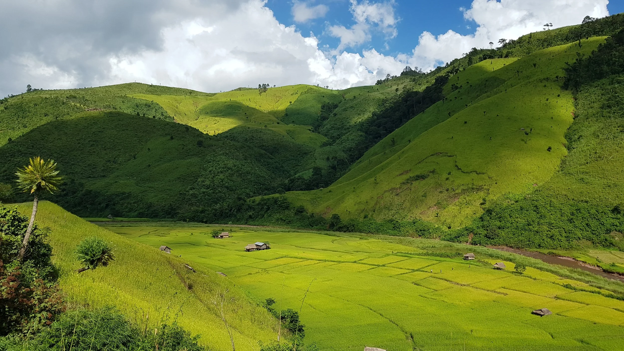 Paysage de collines verdoyantes avec quelques maisons traditionnelles, sous un ciel partiellement nuageux.
