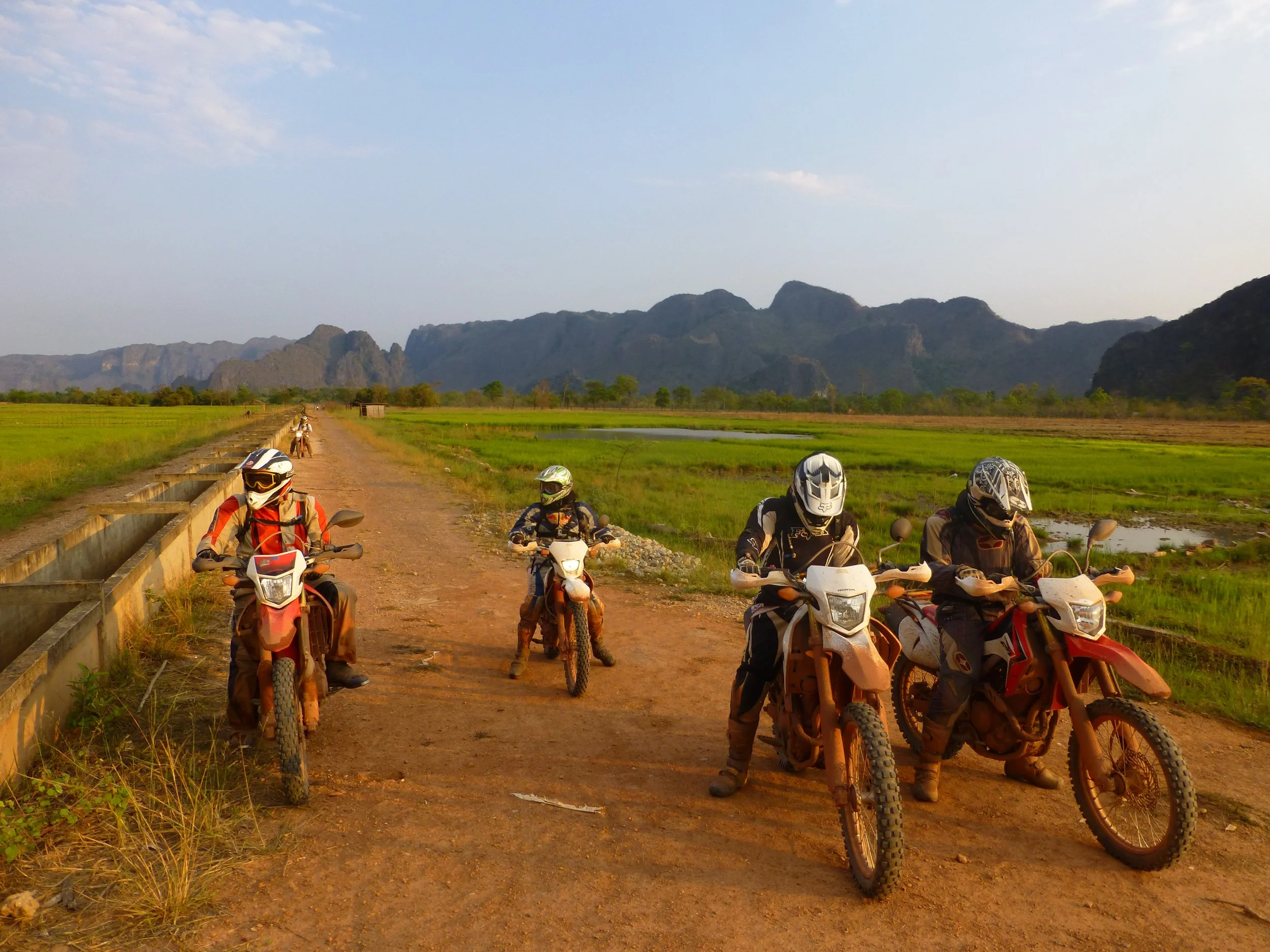 Groupe de motards avec casques en tenue de moto, posant pour une photo devant un paysage rural avec des champs verts et des montagnes en arrière-plan, sous un ciel partiellement nuageux. Au Laos