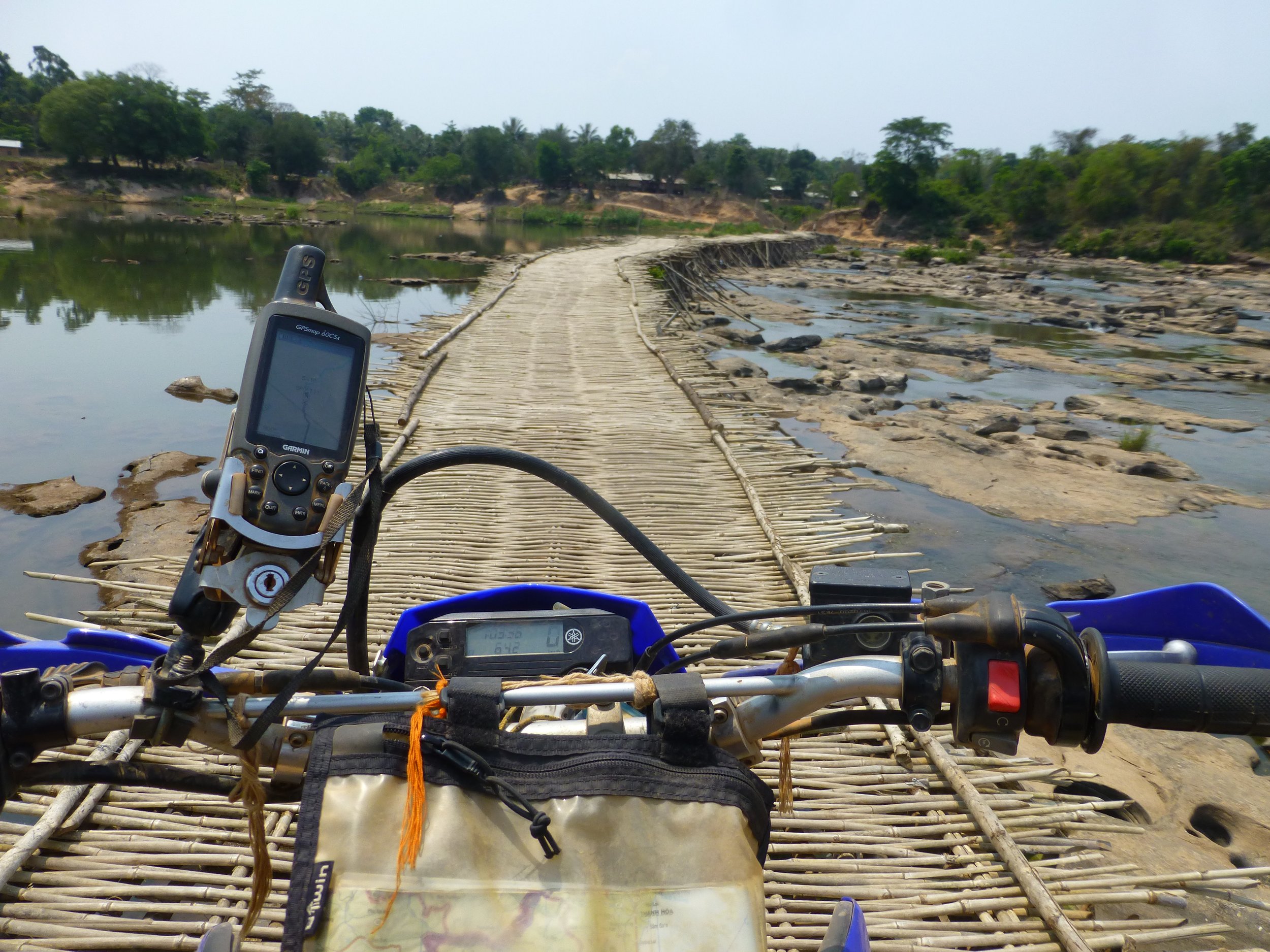 Vue du tableau de bord d'une moto sur un pont en bambou traversant une rivière dans un paysage naturel verdoyant.