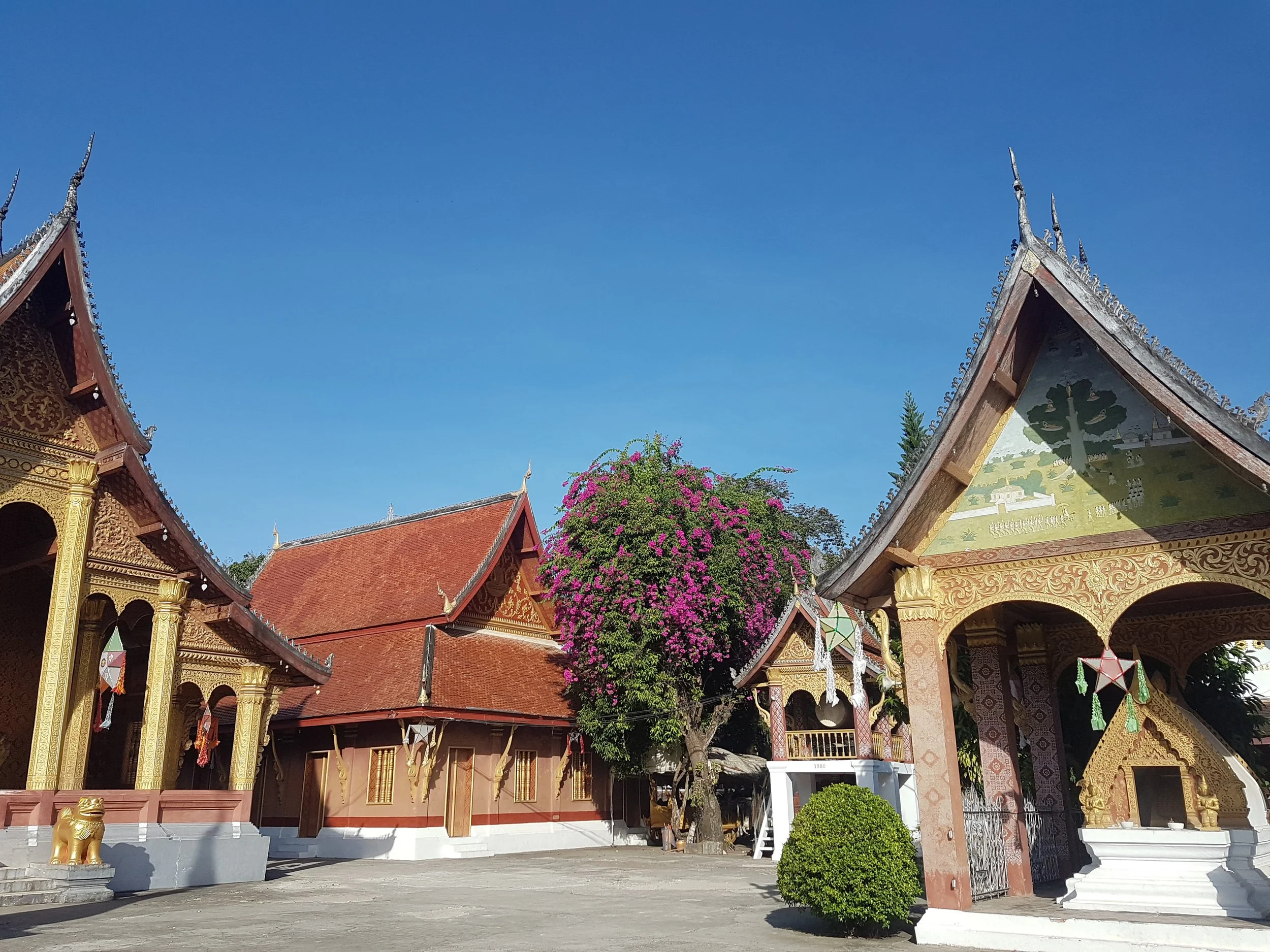 Temples thaïlandais avec toits ornés de sculptures, un arbre fleuri rose et un ciel bleu en arrière-plan.