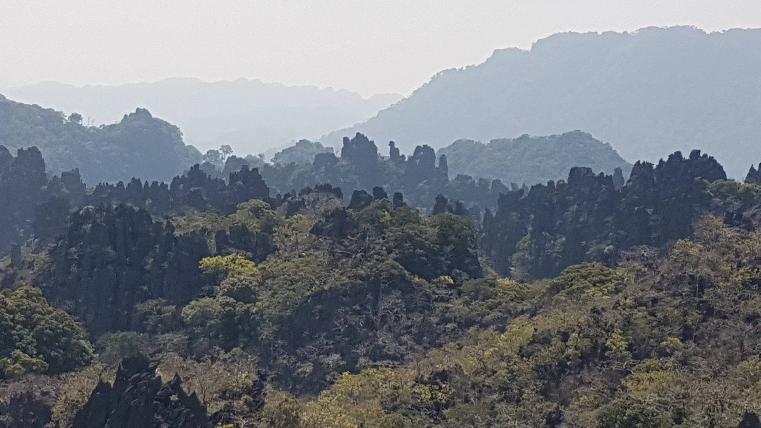 Paysage de montagnes rocheuses couvertes de végétation, avec des sommets en arrière-plan et une teinte brumeuse.