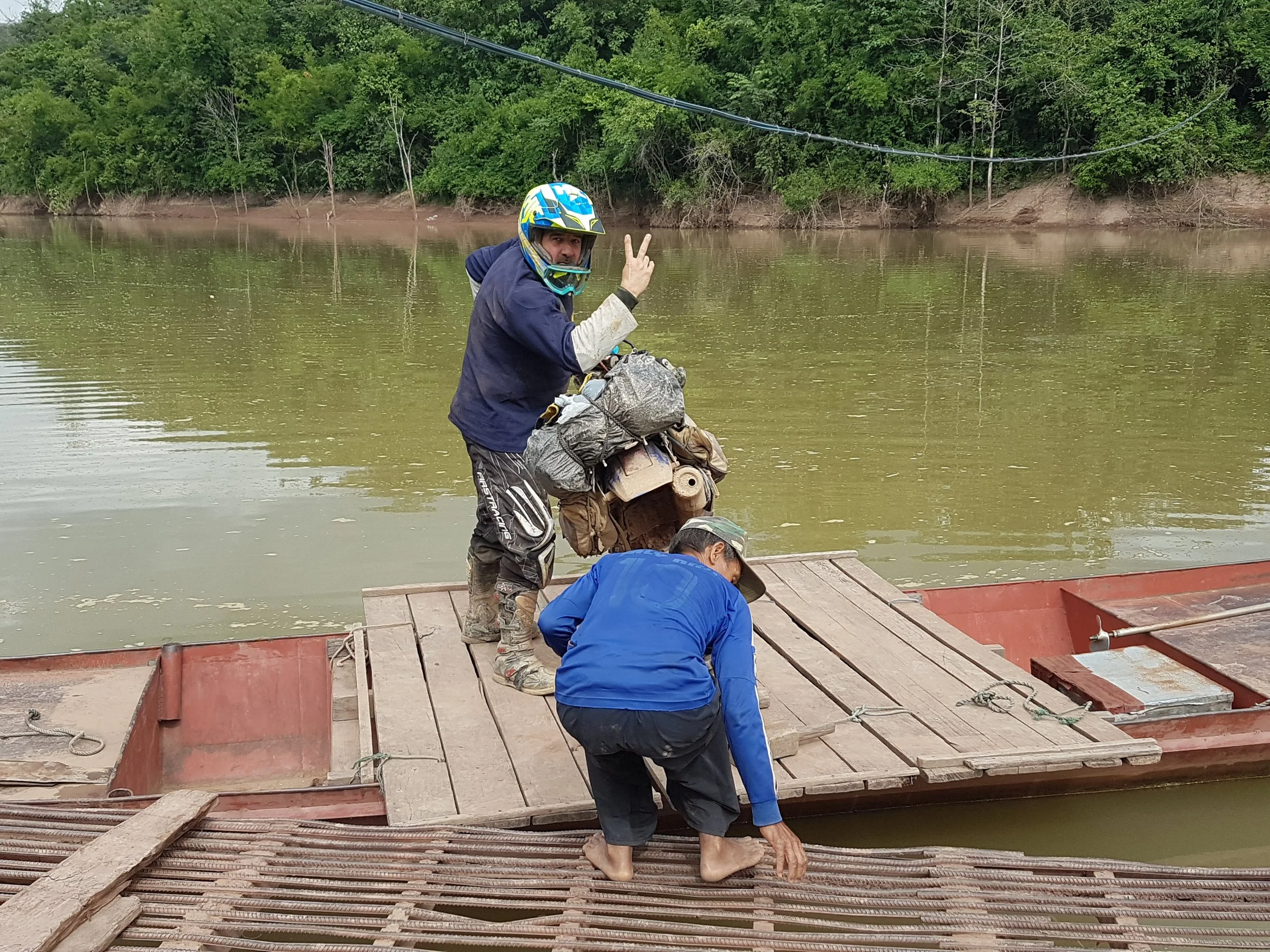 Groupe de personnes sur un pont en bois au bord d'une rivière, portant des équipements de plein air et des casques de sécurité, en train de se préparer à une activité en plein air, avec une forêt dense en arrière-plan.