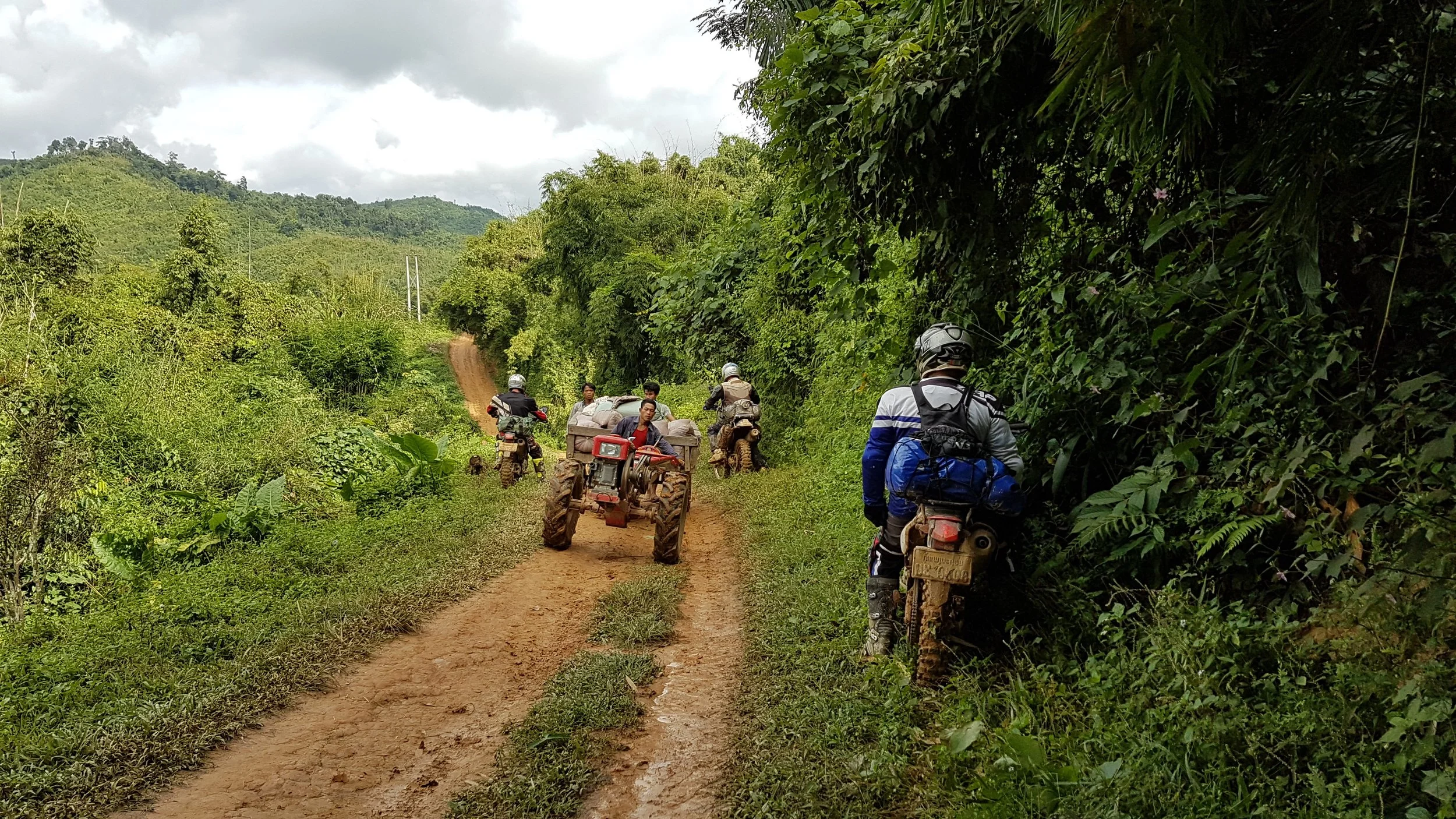 Groupe de personnes avec des motos et une petite moto-tricyle sur un chemin de terre dans une zone forestière dense, montagne en arrière-plan.