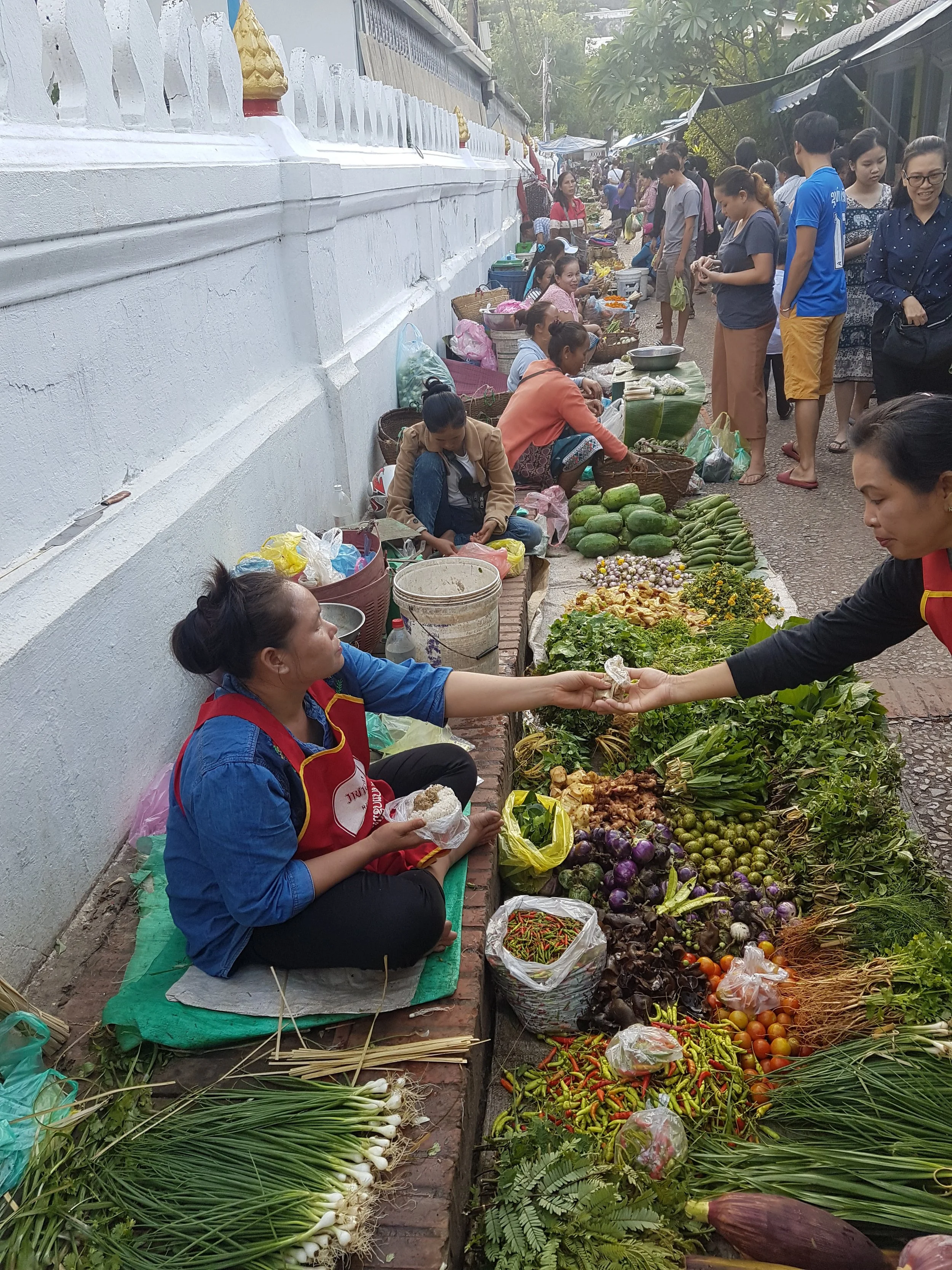 Marché en plein air avec des vendeurs vendant des légumes et des fruits, et des clients achetant, le tout dans une rue pavée bordée d'une cloison blanche décorée.