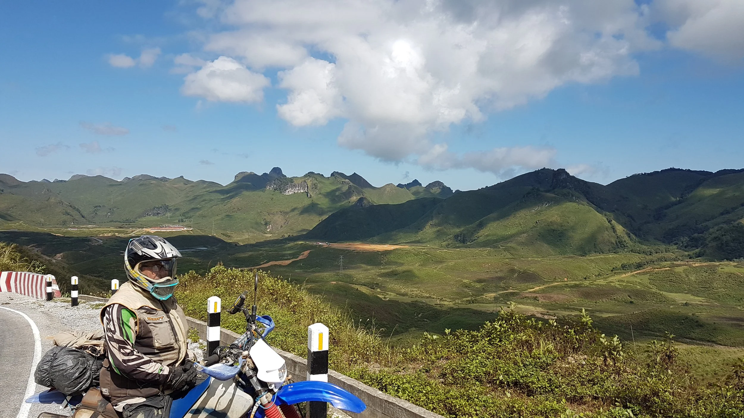 Un motard portant un casque et une veste de moto, avec une sacoche sur le côté, se trouve à côté de sa moto bleue. En arrière-plan, on voit un paysage de montagnes verdoyantes sous un ciel partiellement nuageux.