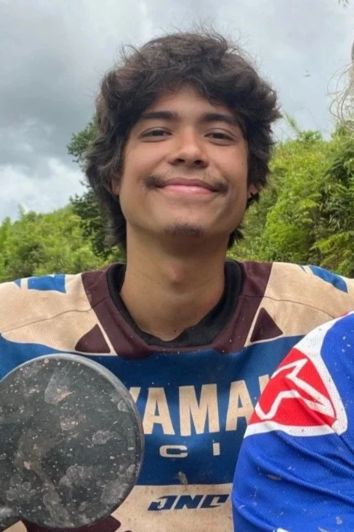 Un jeune homme souriant avec des cheveux bouclés, portant un maillot de sport rayé de beige, bleu et blanc, à l'extérieur avec des arbres en arrière-plan.