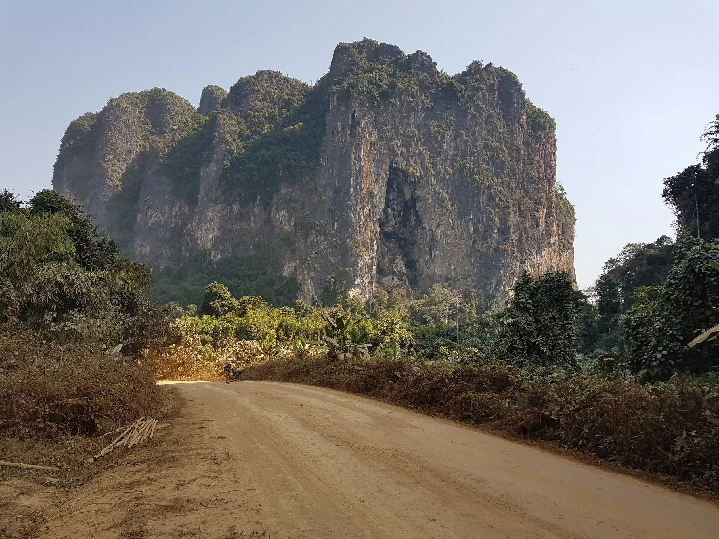 Une route en terre entourée de végétation dense menant à une montagne rocheuse impressionnante avec des formations escarpées et de la verdure sur ses sommets, sous un ciel clair.