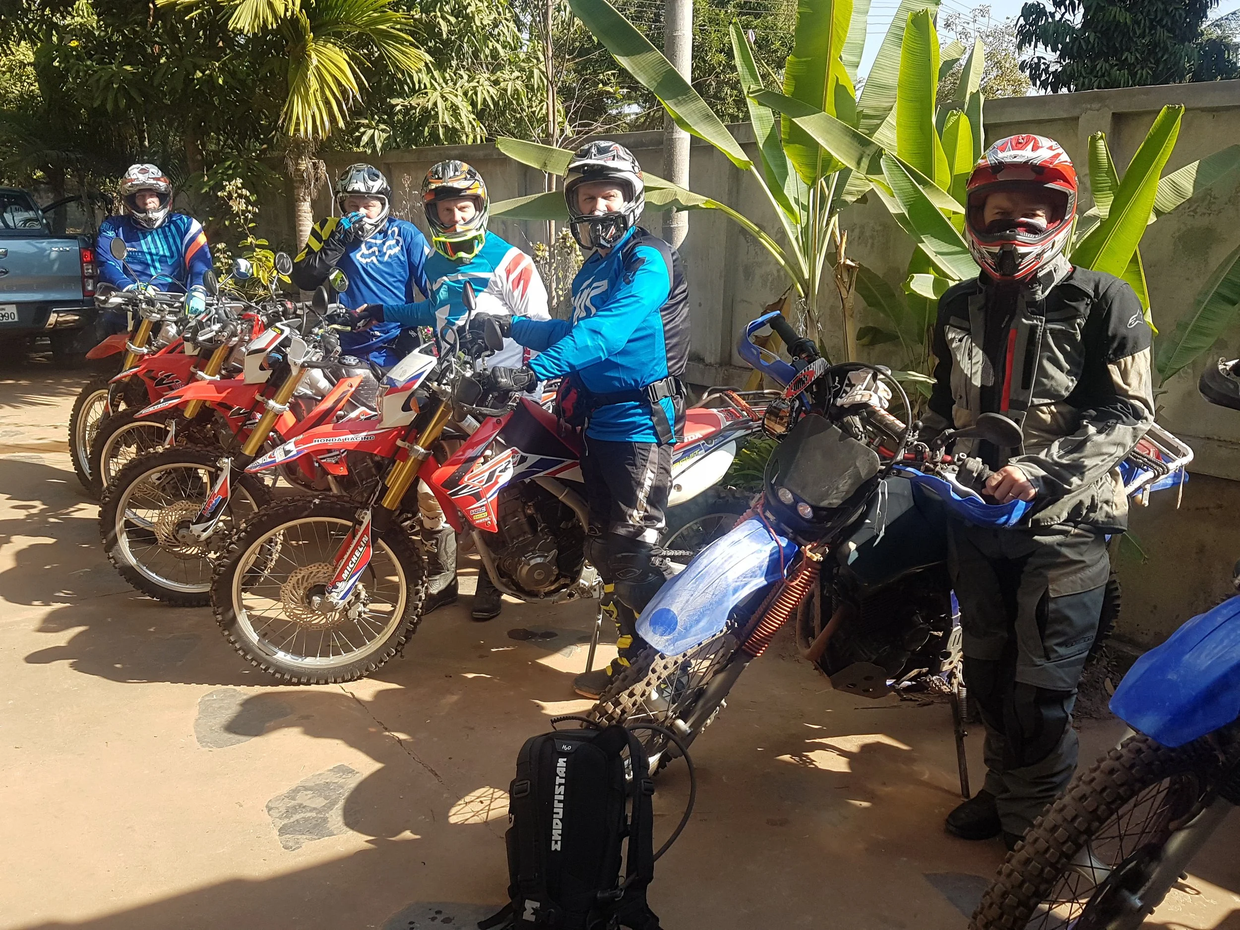 Groupe de cinq personnes en tenues de motocross, portant des casques, posant avec des motos tout-terrain rouges et bleues, à l'extérieur sous un clair soleil, entouré de végétation tropicale.