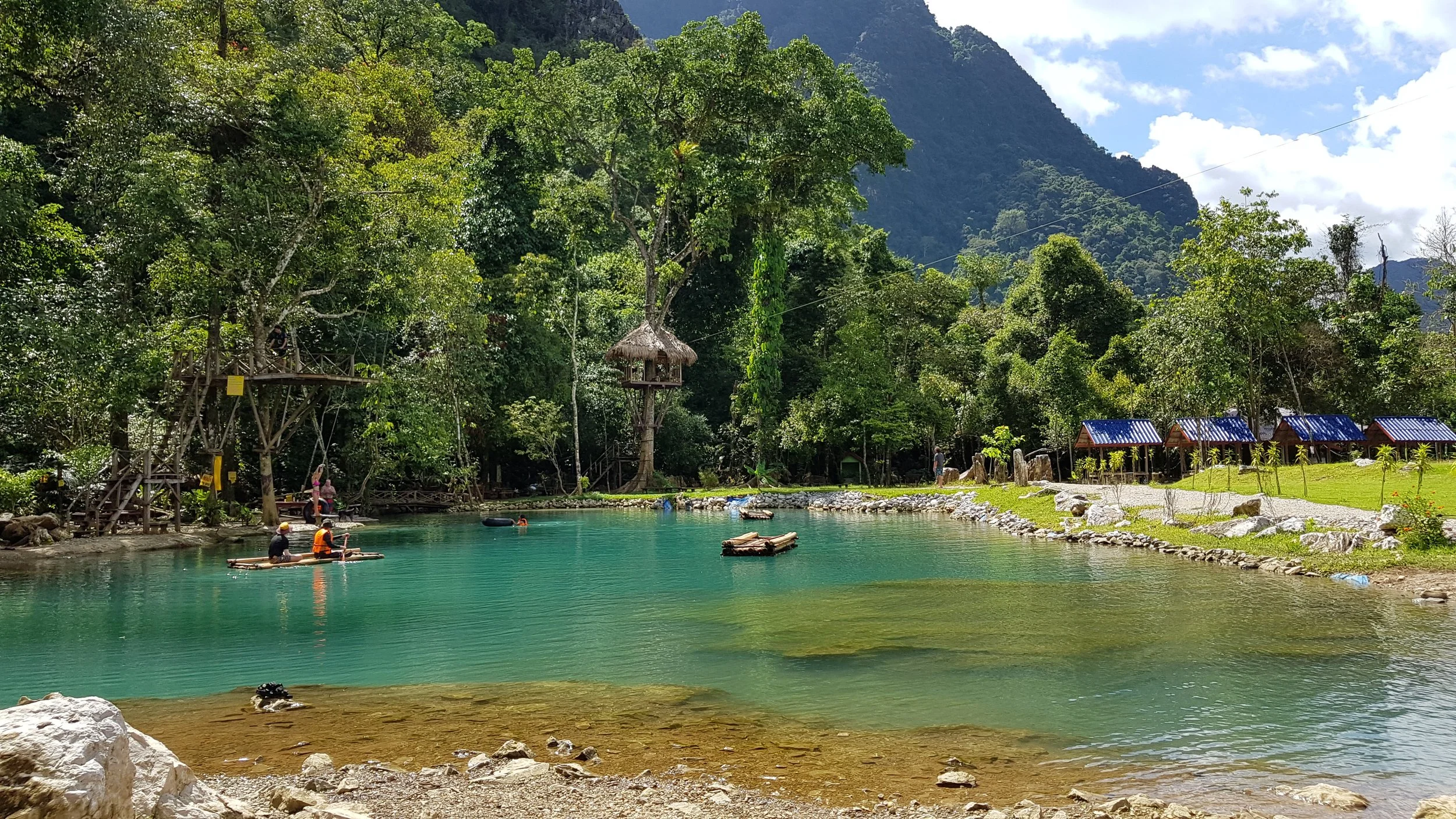 Lac turquoise entouré de nature verdoyante avec des montagnes en arrière-plan, des huttes en bois, et des personnes en train de faire du paddle sur l'eau.