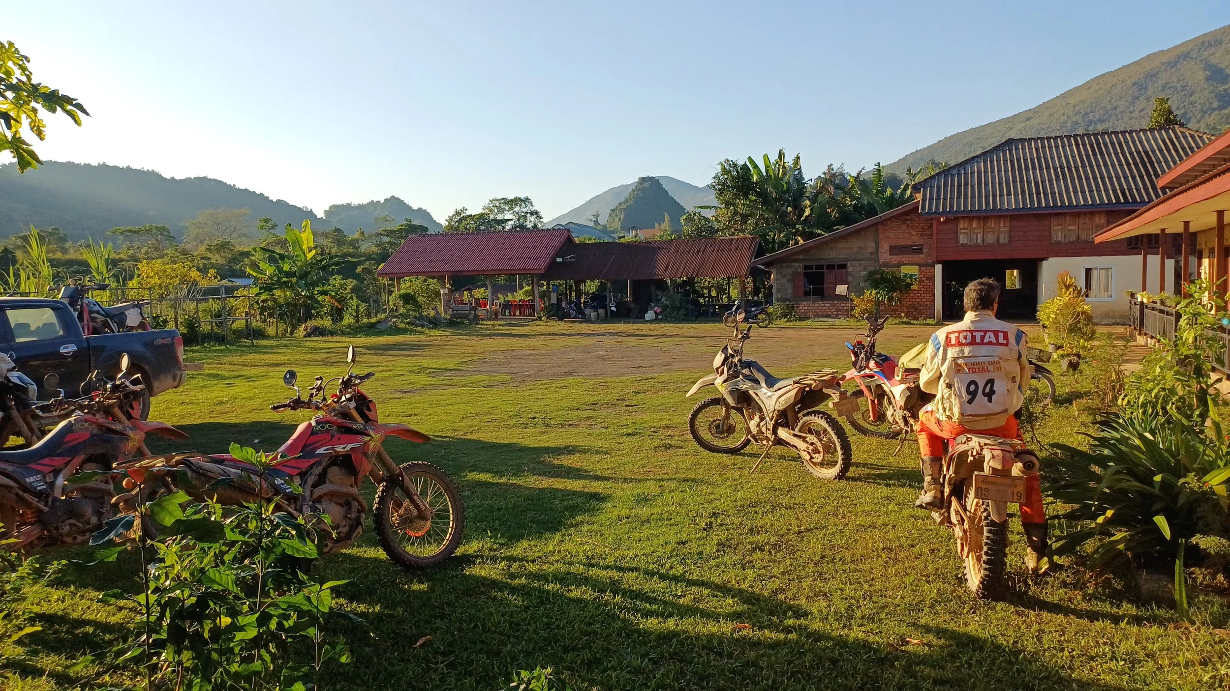 Un homme en veste avec le logo TOTAL assis sur une moto en plein air, avec plusieurs motos et un bâtiment en arrière-plan, entouré de montagne et de verdure.