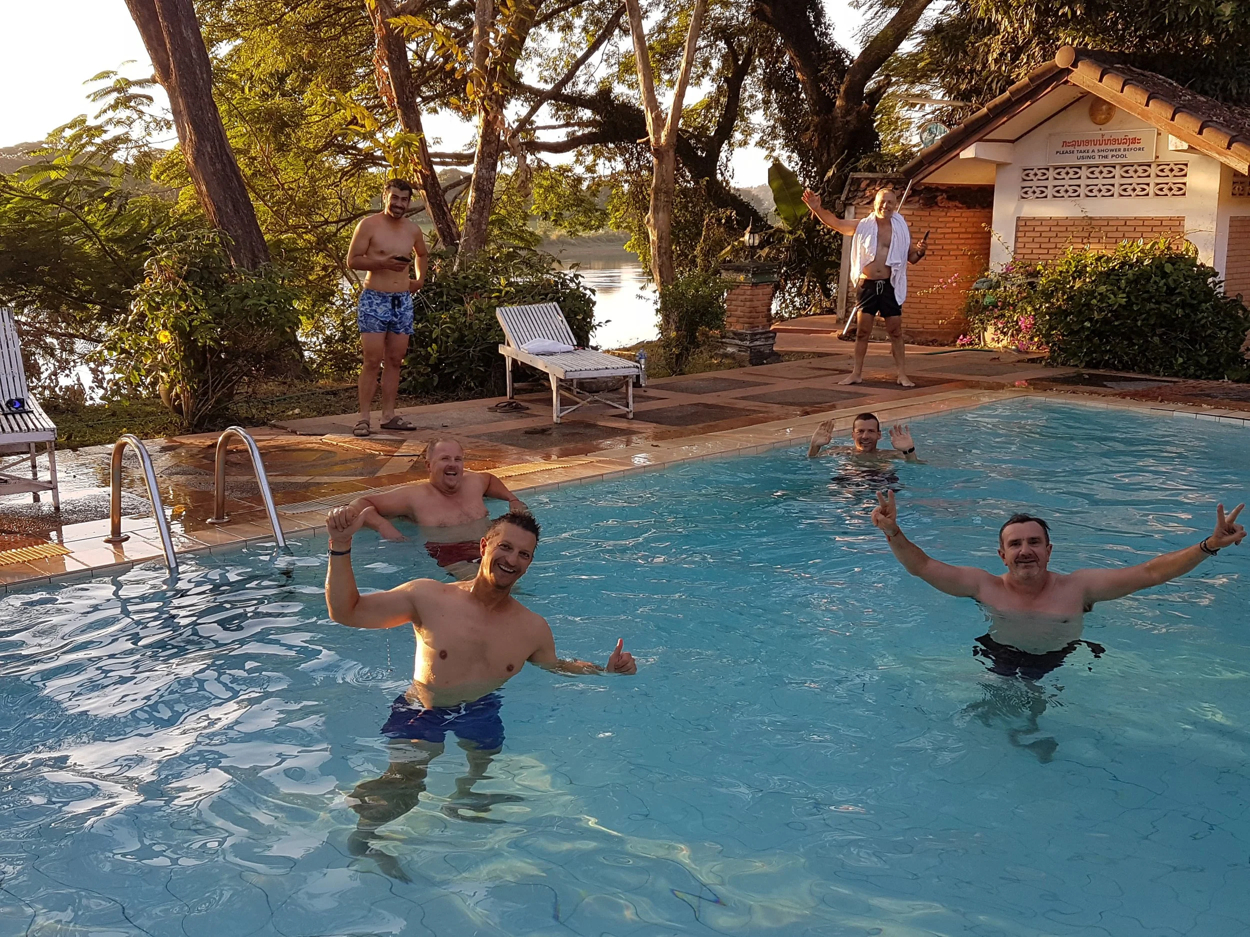 Groupe d'hommes nageant et souriant dans une piscine en plein air avec un paysage naturel en arrière-plan, en fin de journée