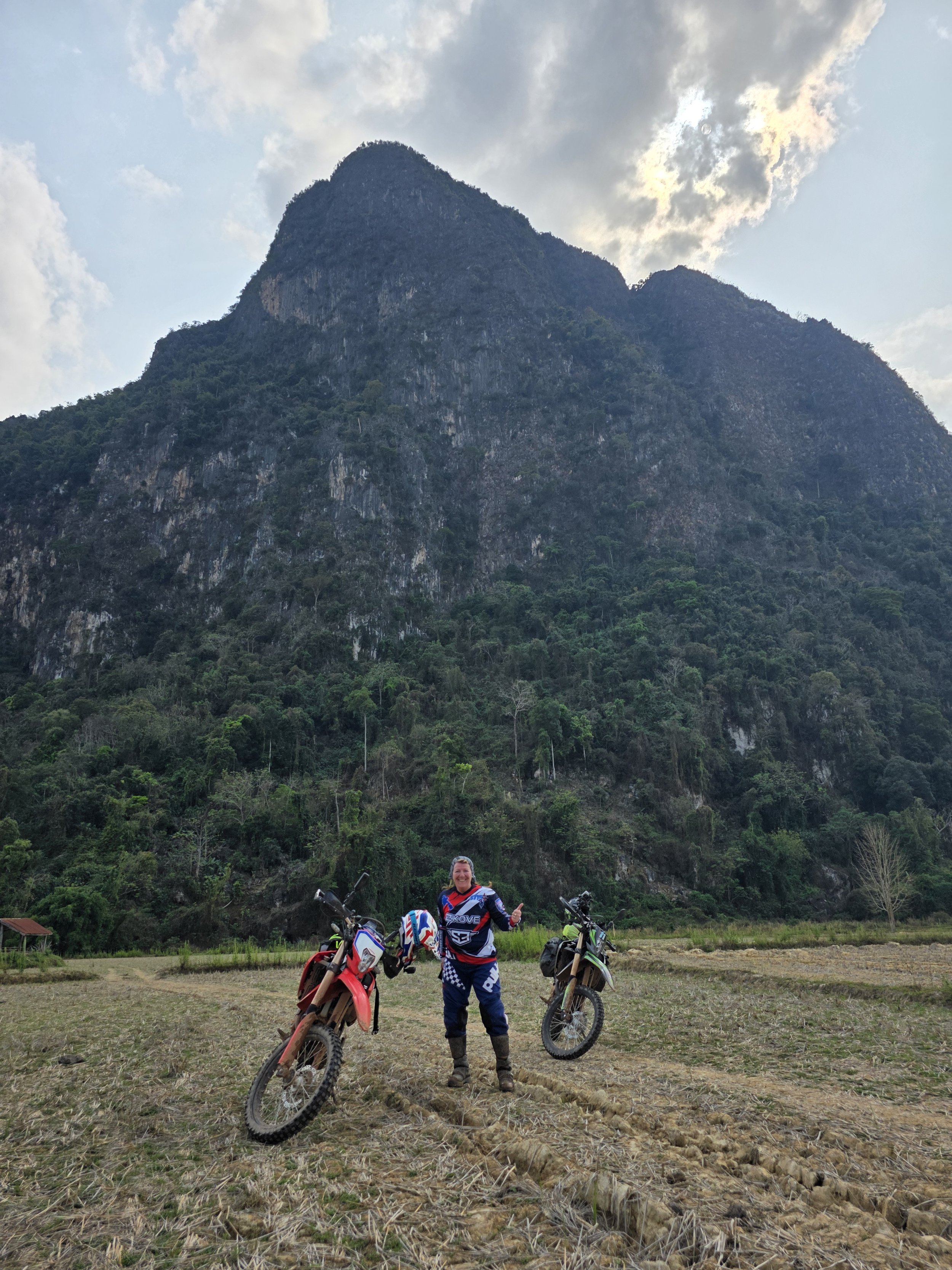 Une femme en équipement de moto tout-terrain, souriante et faisant un signe de pouce en l'air, se tient entre trois motos tout-terrain dans un champ avec une montagne en arrière-plan et un ciel partiellement nuageux.