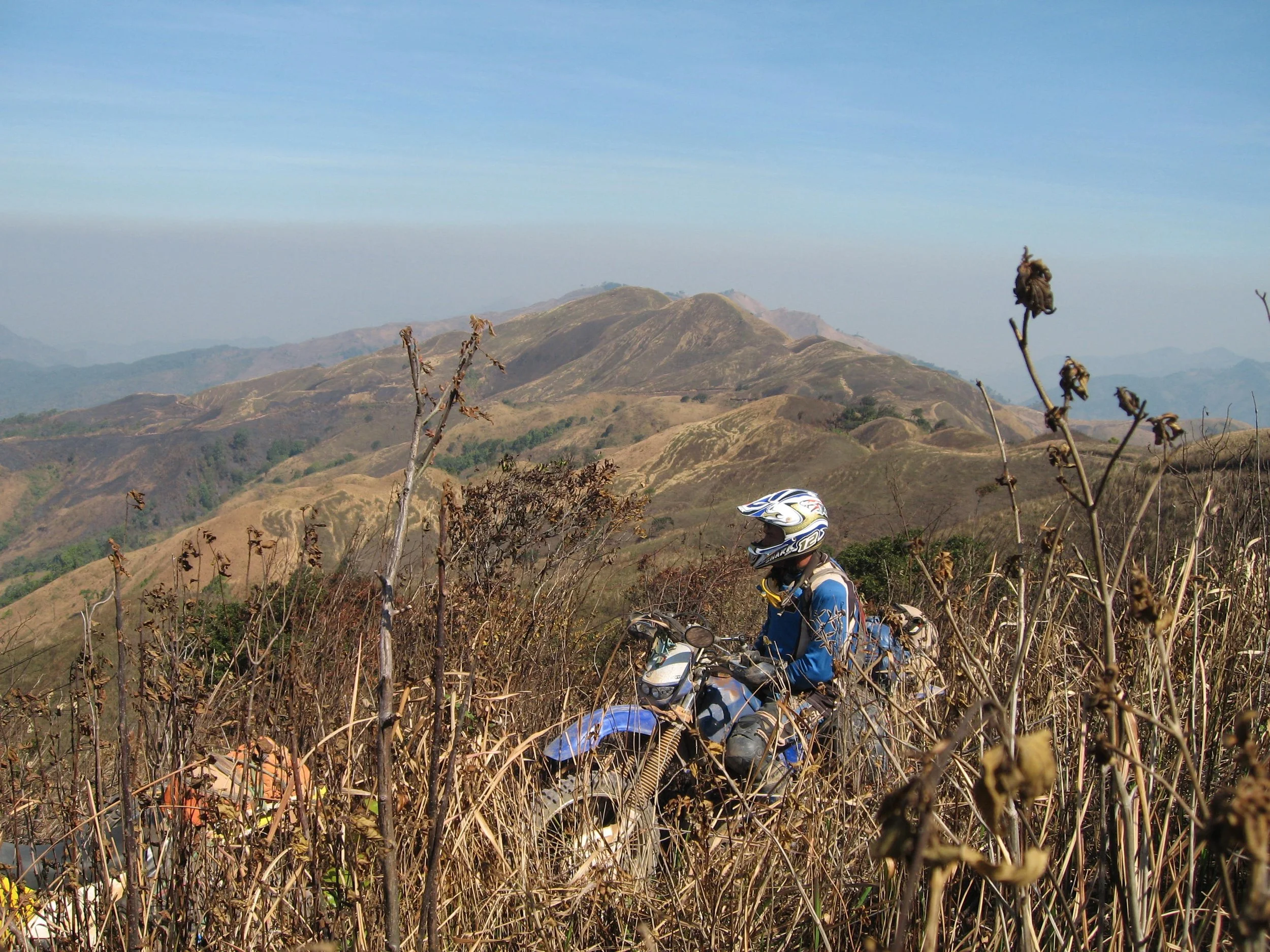 Un paysage montagneux avec des collines et des montagnes sous un ciel clair au Laos. Un motard avec un casque et une veste bleue est visible parmi des branches sèches et des végétaux au premier plan.