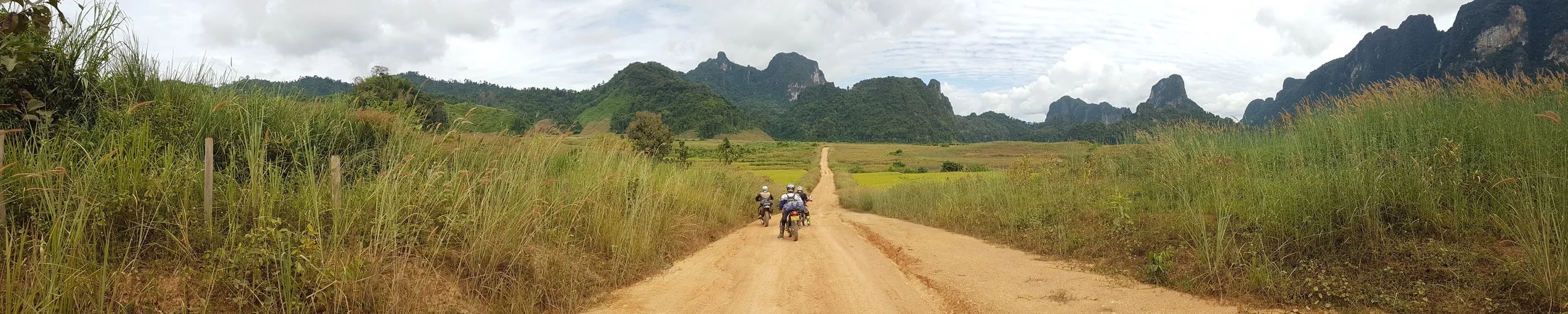 Paysage rural avec une piste en terre, montagnes en arrière-plan, et quatre motos roulant sur la piste.