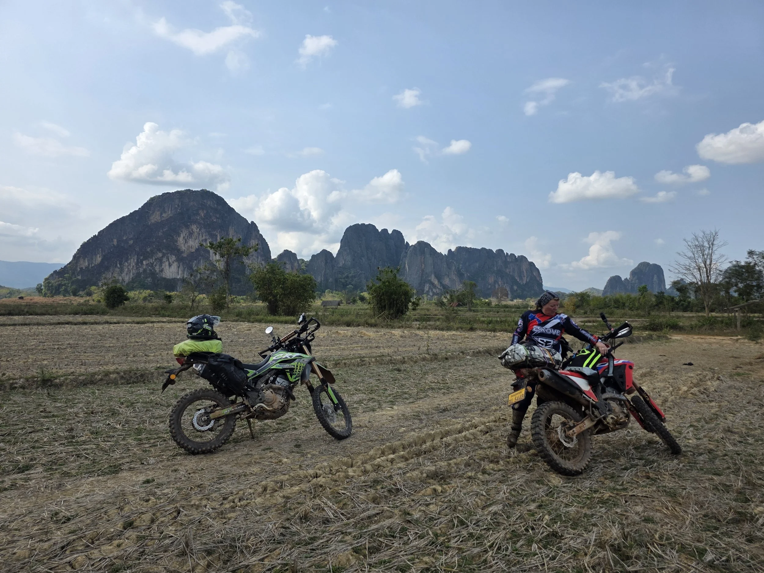 Deux motards dans un champ avec des montagnes en arrière-plan, ciel partiellement nuageux.